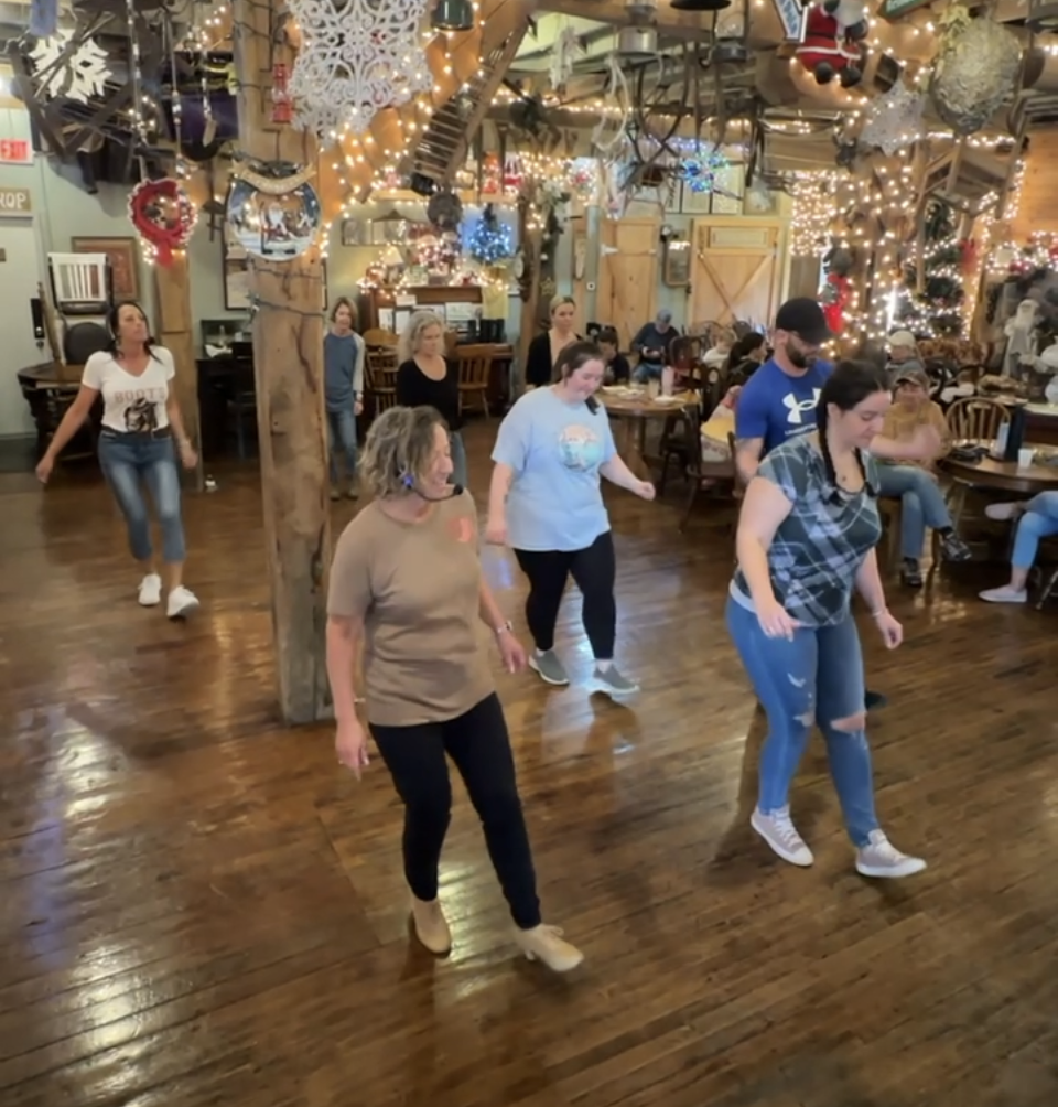 People line dancing in a decorated rustic restaurant with Christmas lights and ornaments.
