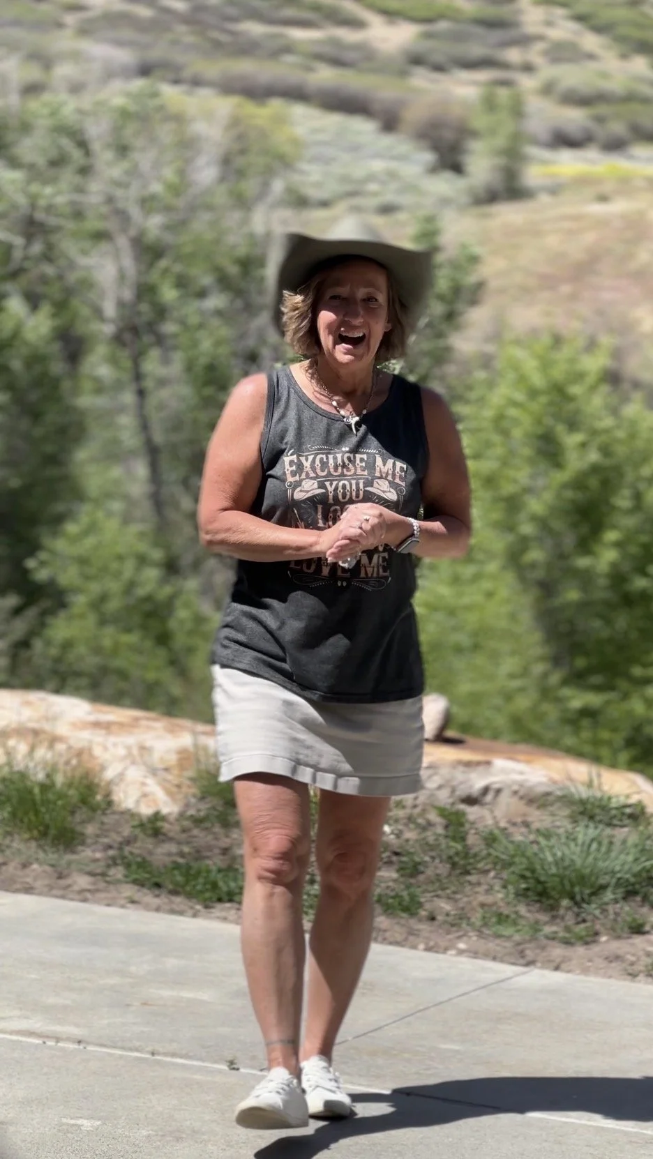 A woman smiling and standing outdoors on a sunny day, wearing a cowboy hat, black sleeveless shirt, and light-colored shorts with a scenic natural background of trees and hills.
