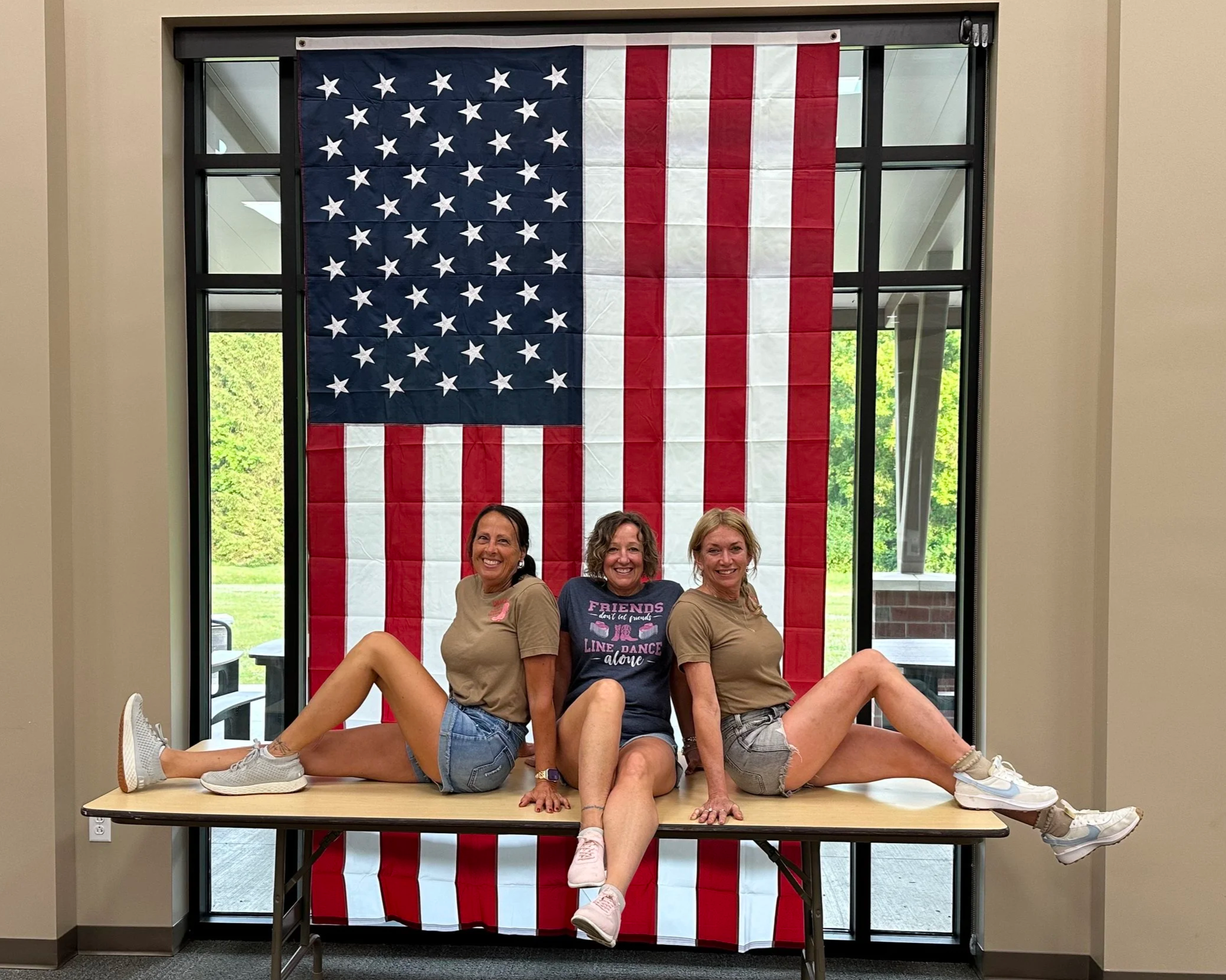 Three women sitting on a table with their legs extended and intertwined, posing in front of an American flag hanging on a window behind them.