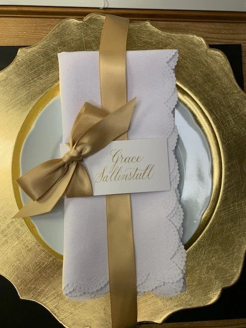 Wedding place card written in calligraphy full names in gold ink with attached gold satin ribbon and bow at place setting for wedding decor.