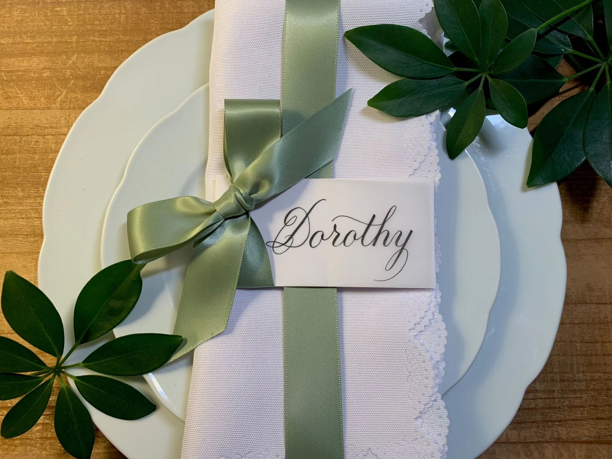 Calligraphy place card decorated with a sage satin ribbon and tied in a bow shown at a table setting at wedding event.