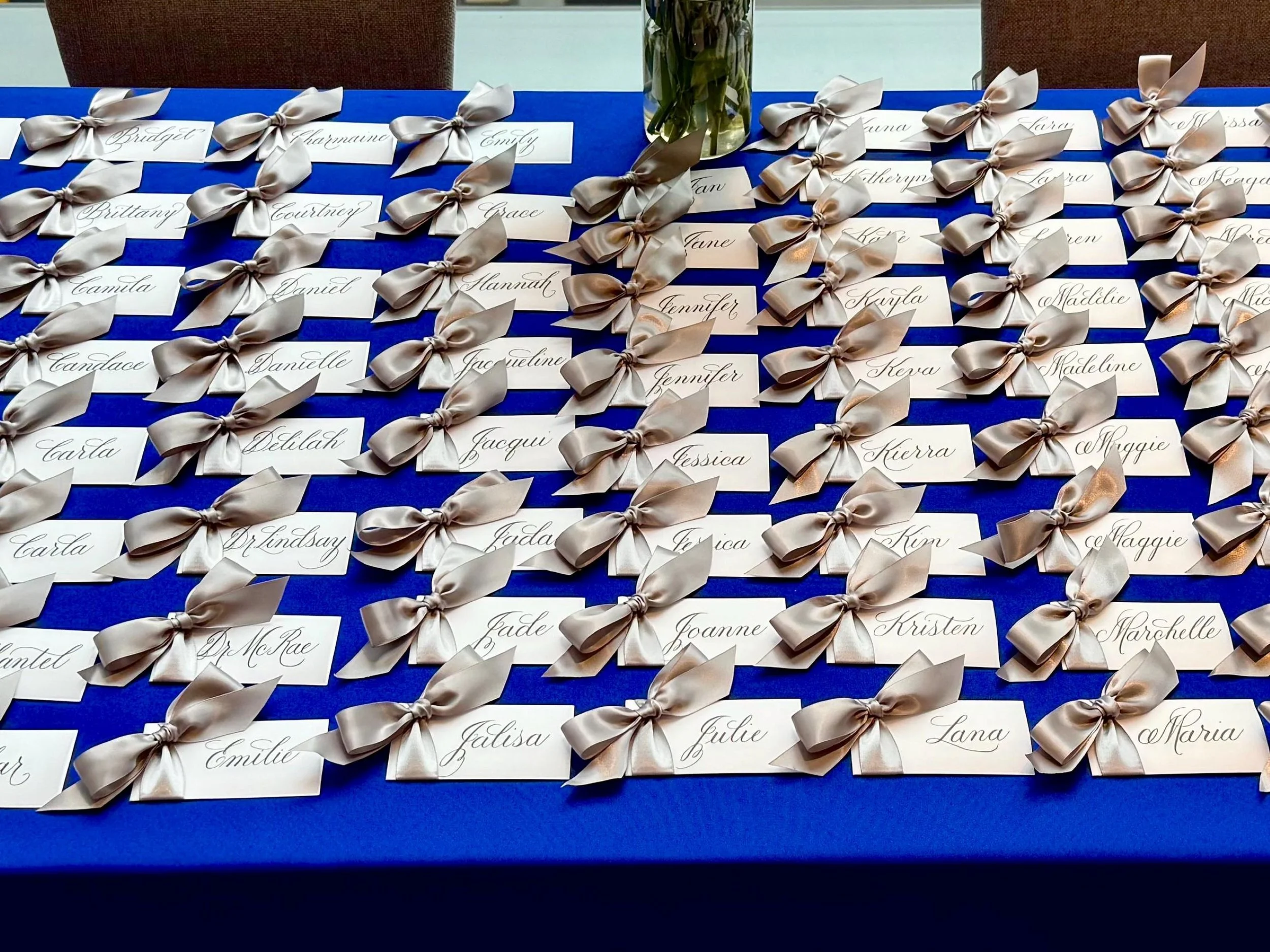 Handwritten calligraphy name tags with elegant writing and silver satin bows arranged on a royal blue tablecloth at a business conference.