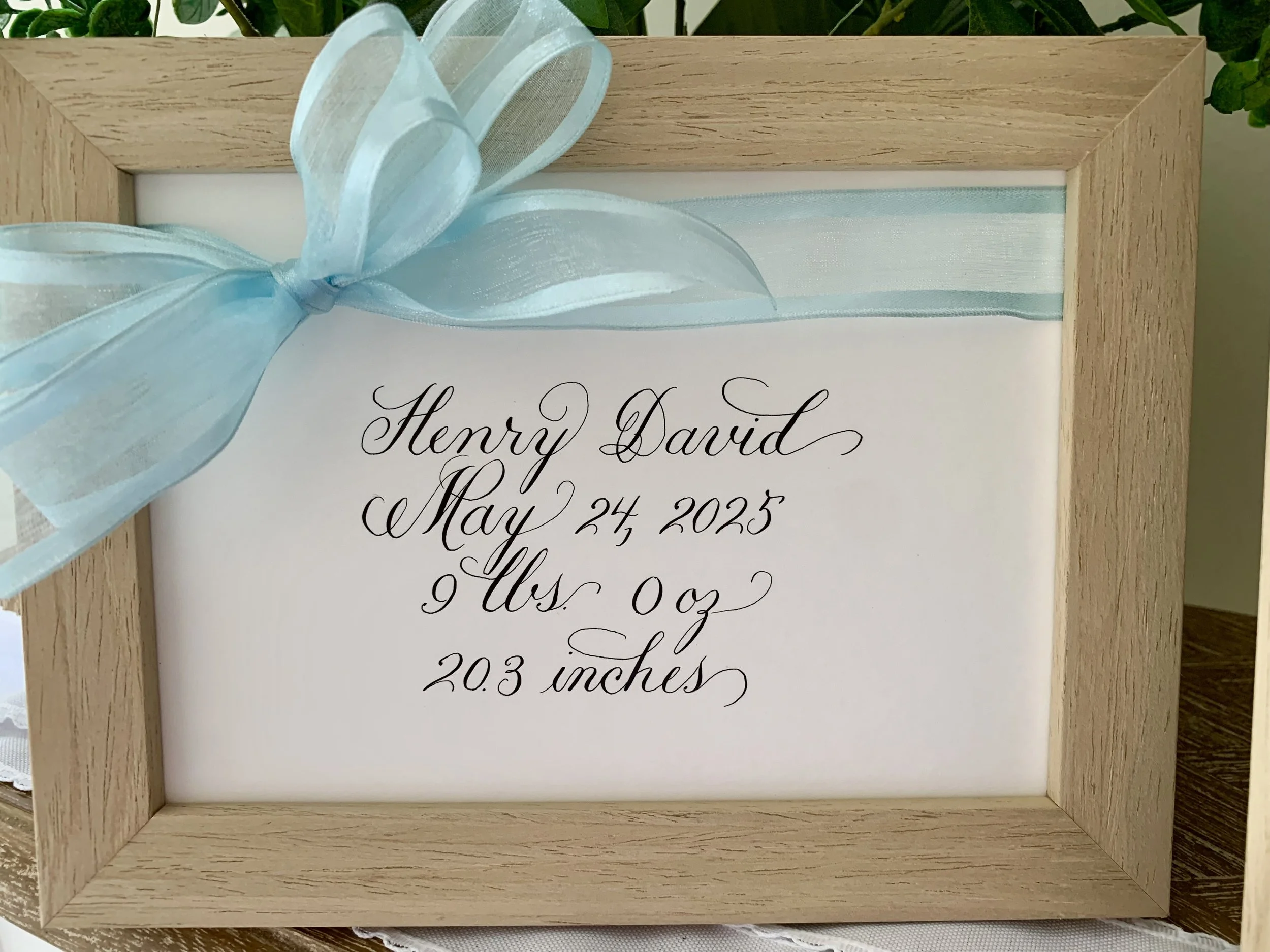 Baby announcement gift of framed calligraphy baby name, date of birth, and weight deocrated with blue ribbon and bow for nursery decor.