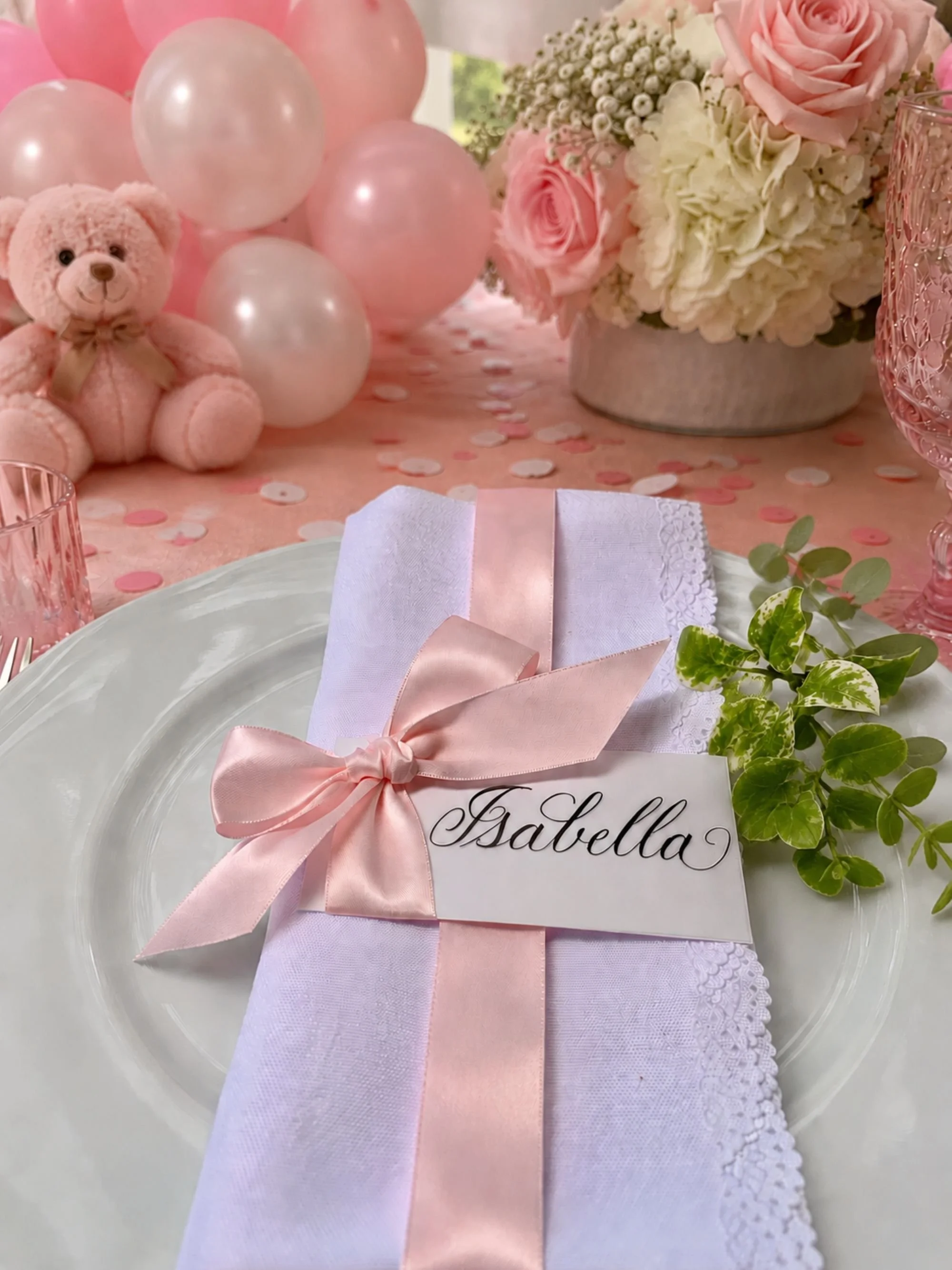 Calligraphy pink ribbon place card with baby pink satin ribbon tied in a bow laying on a napkin and plate on a table decorated with balloons and teddy bear at a baby shower.