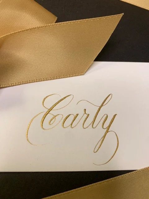 Elegant gold ribbon place card handwritten in gold ink calligraphy for wedding or party.