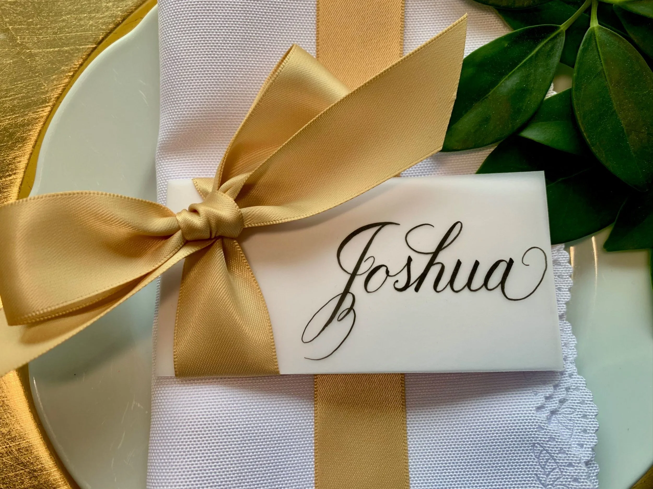 Gold satin ribbon and bow place card with handwritten calligraphy name placed on plate at event
