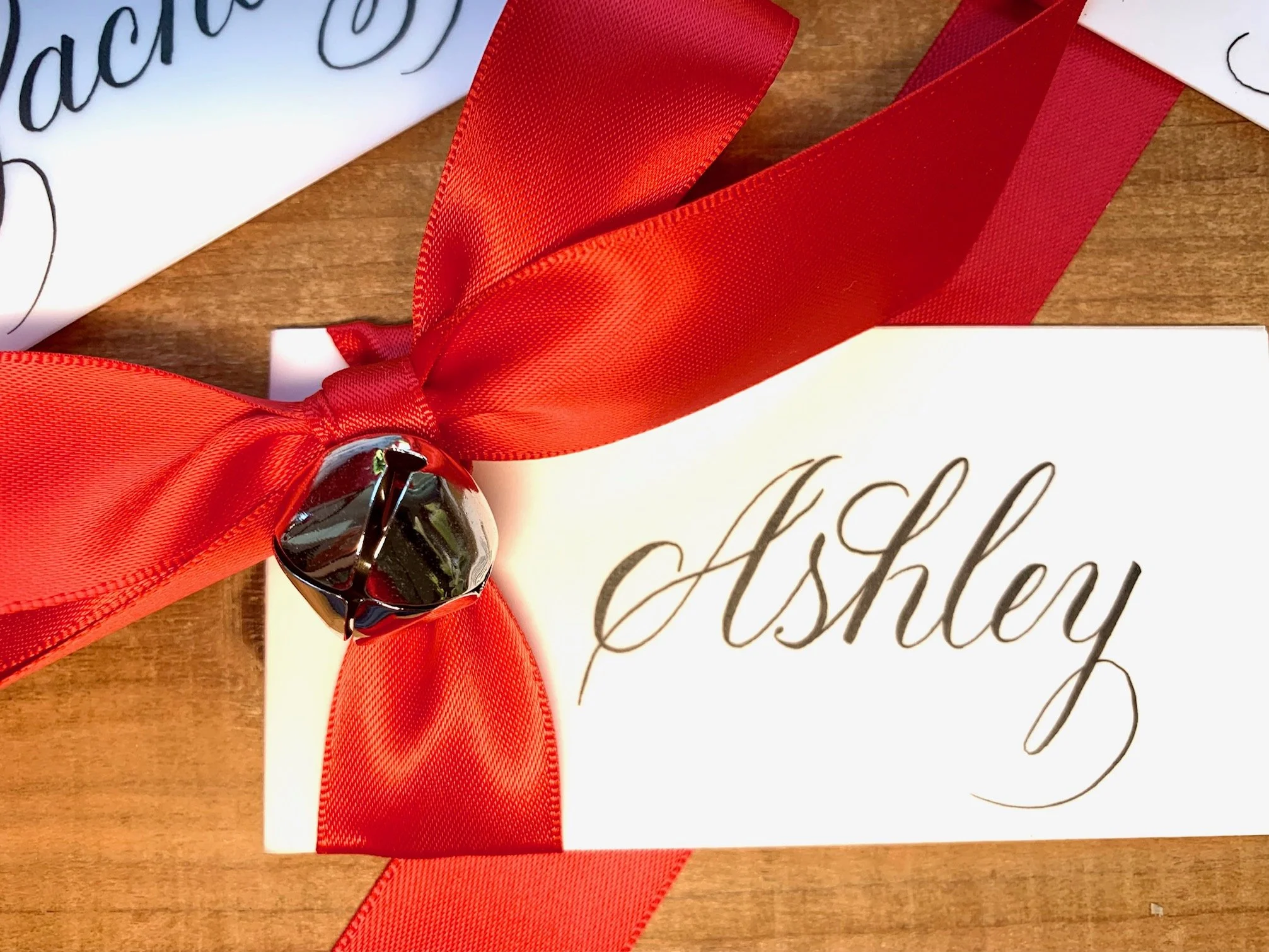 Calligraphy Christmas place card with handwritten first name and decorated with jingle bell and red satin bow