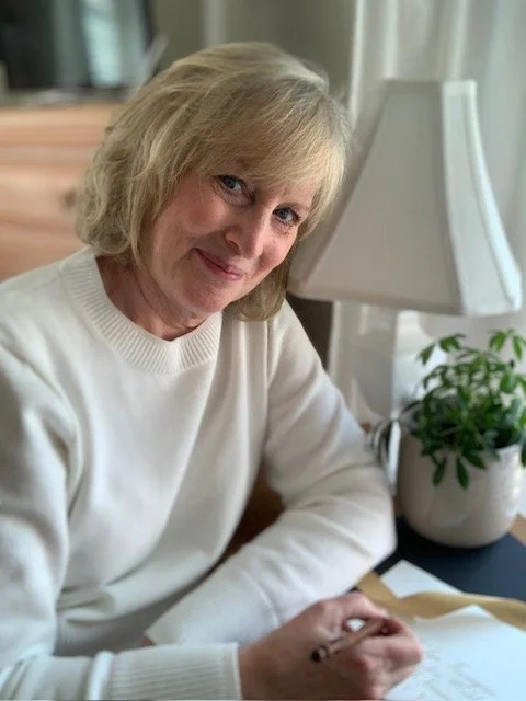 A woman with shoulder-length blonde hair smiling and seated at a desk with papers, beside a table lamp and a potted plant.
