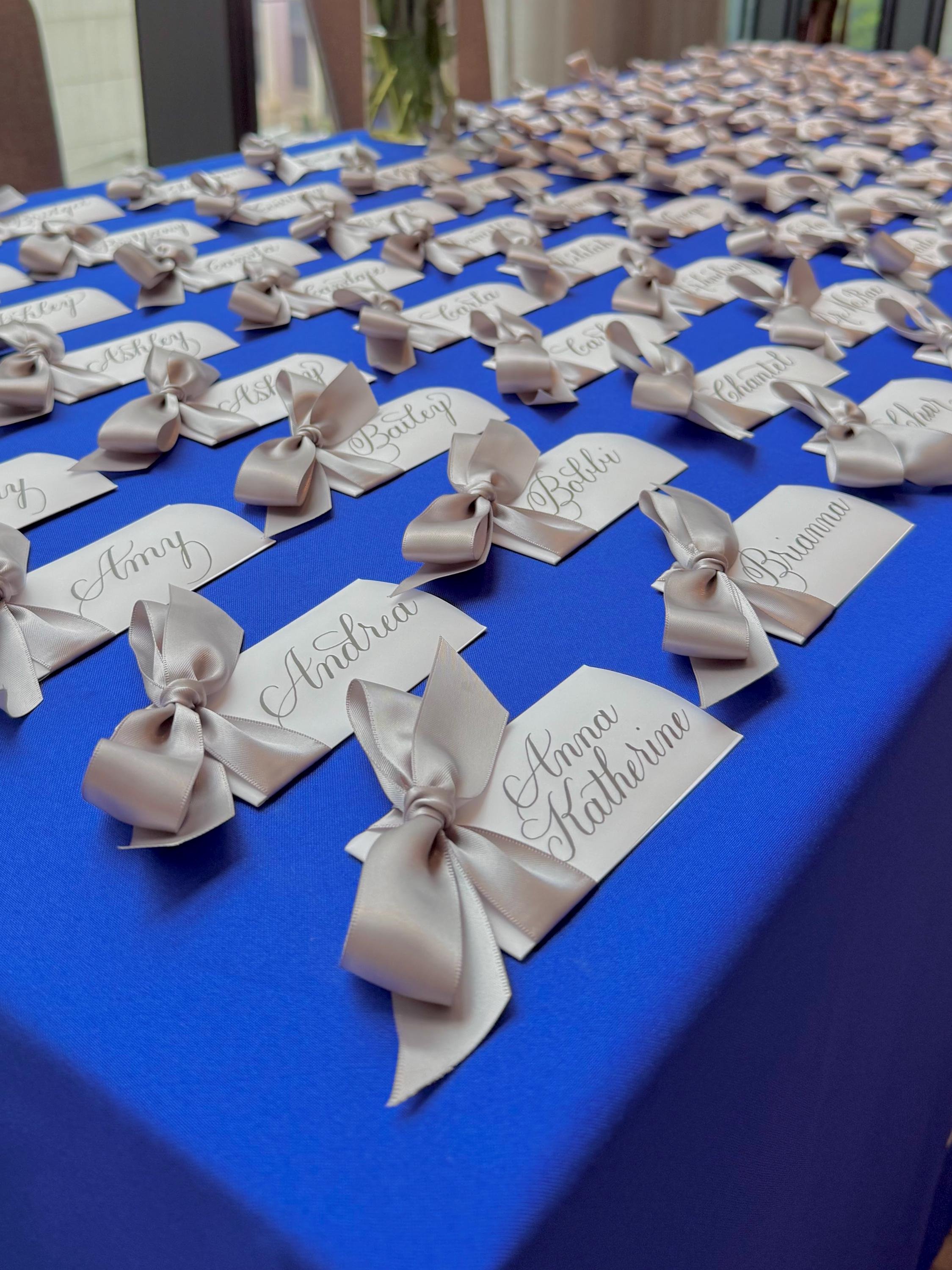 Table with white name cards decorated with silver bows, all with handwritten calligraphy names, on a royal blue tablecloth at a business event.