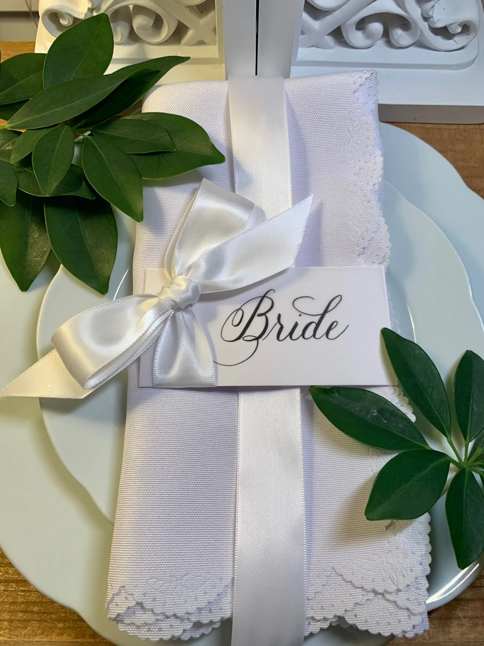 "Bride" placecard handwritten in calligraphy  with attached white satin ribbon and bow laying on place setting at reception.
