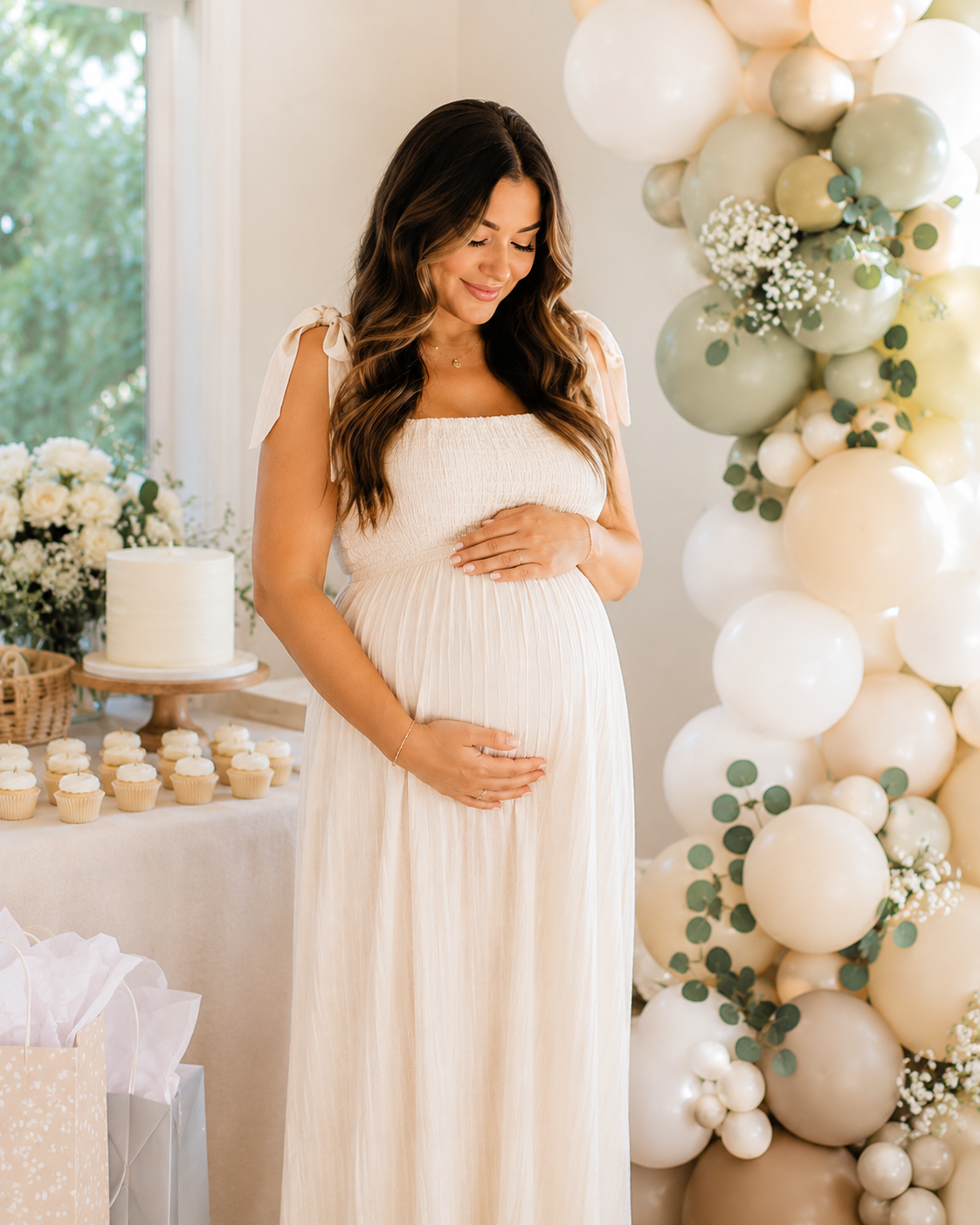 A pregnant woman in a cream-colored dress gently holding her belly with both hands, standing in front of a balloon arrangement, cupcakes, and cake at a baby shower event.