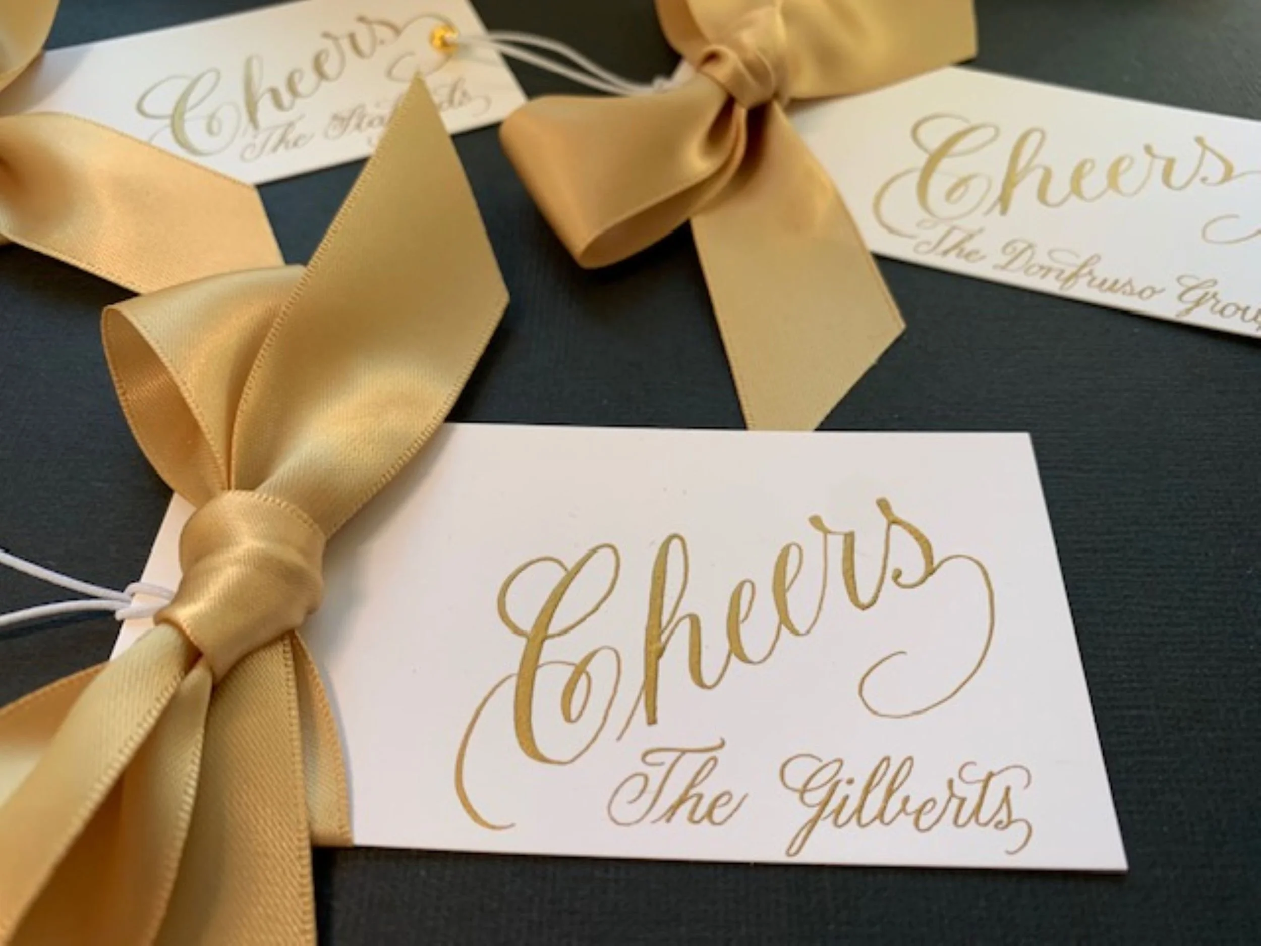 Wine bottle gift tag with personalized gold ink calligraphy of word Cheers and name with gold satin ribbon and bow.