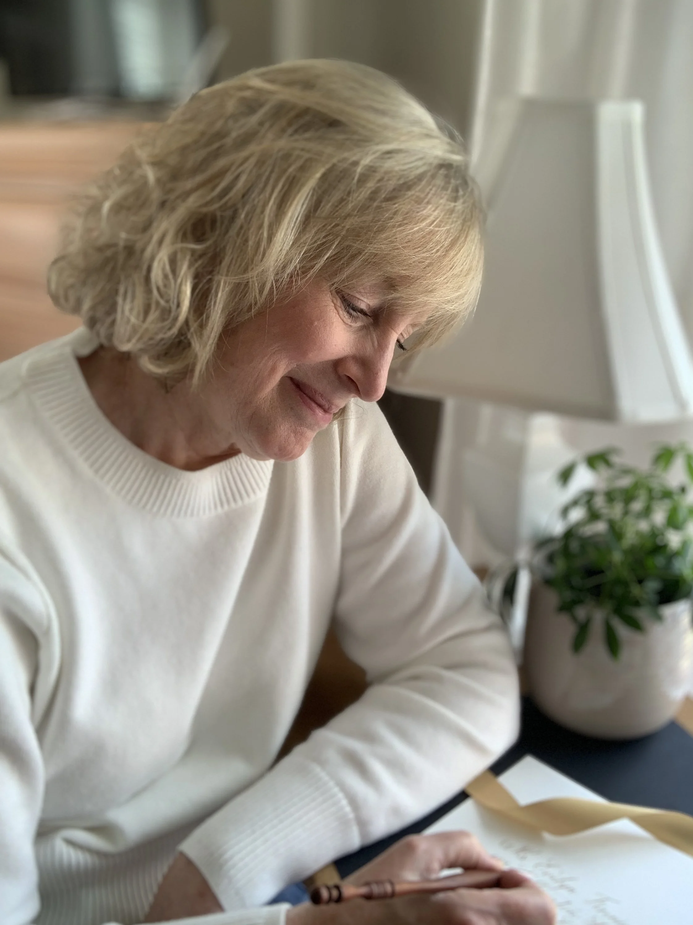 A woman with blonde curly hair wearing a white sweater is writing in a notebook with a brown pen. There is a houseplant with green leaves in the background near a window.