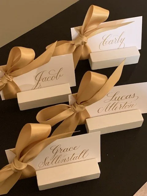 Gold ink place cards with attached gold satin ribbon and bow with first names or full names displayed in card holders.