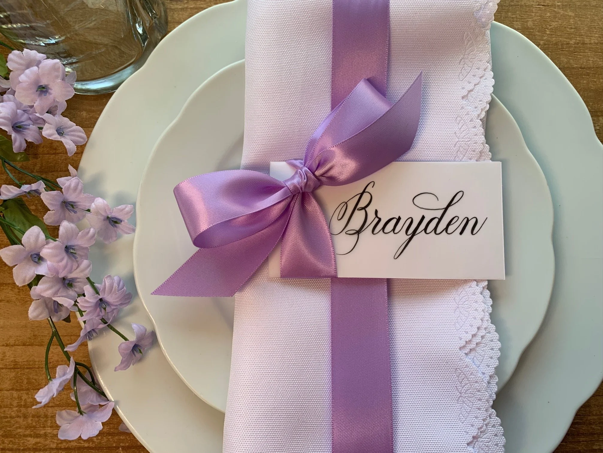 Lavender ribbon-embellished place card with bow and calligraphy name laying on a place setting at party event.
