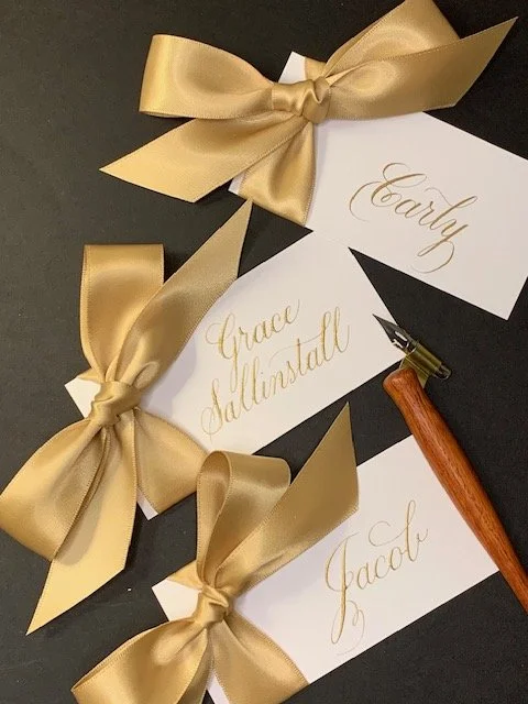 Gold satin bow calligraphy place cards with names handwritten in gold ink for wedding or anniversary party.