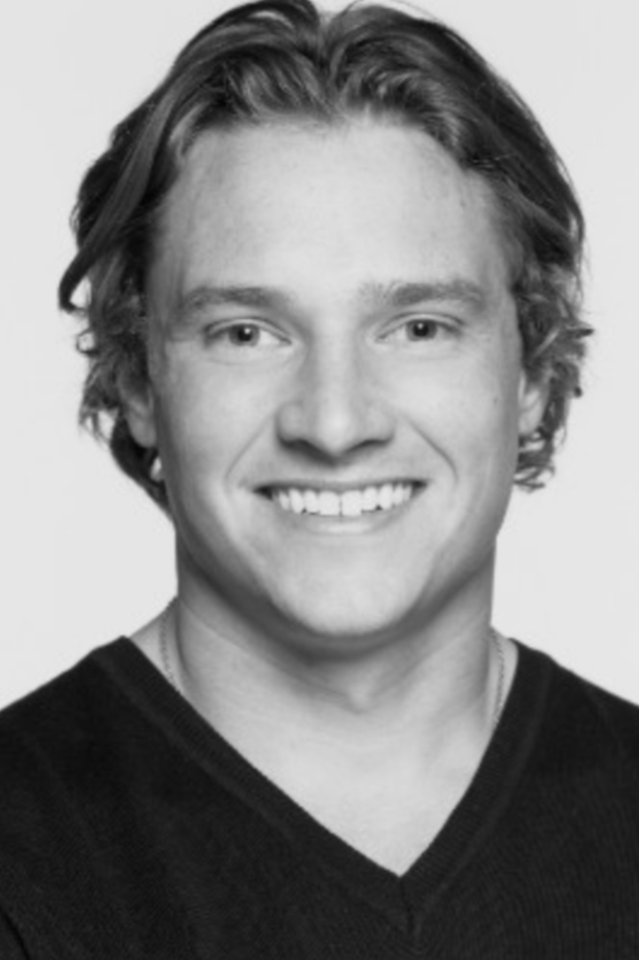 Black and white headshot of a young man with wavy hair smiling, wearing a V-neck sweater.