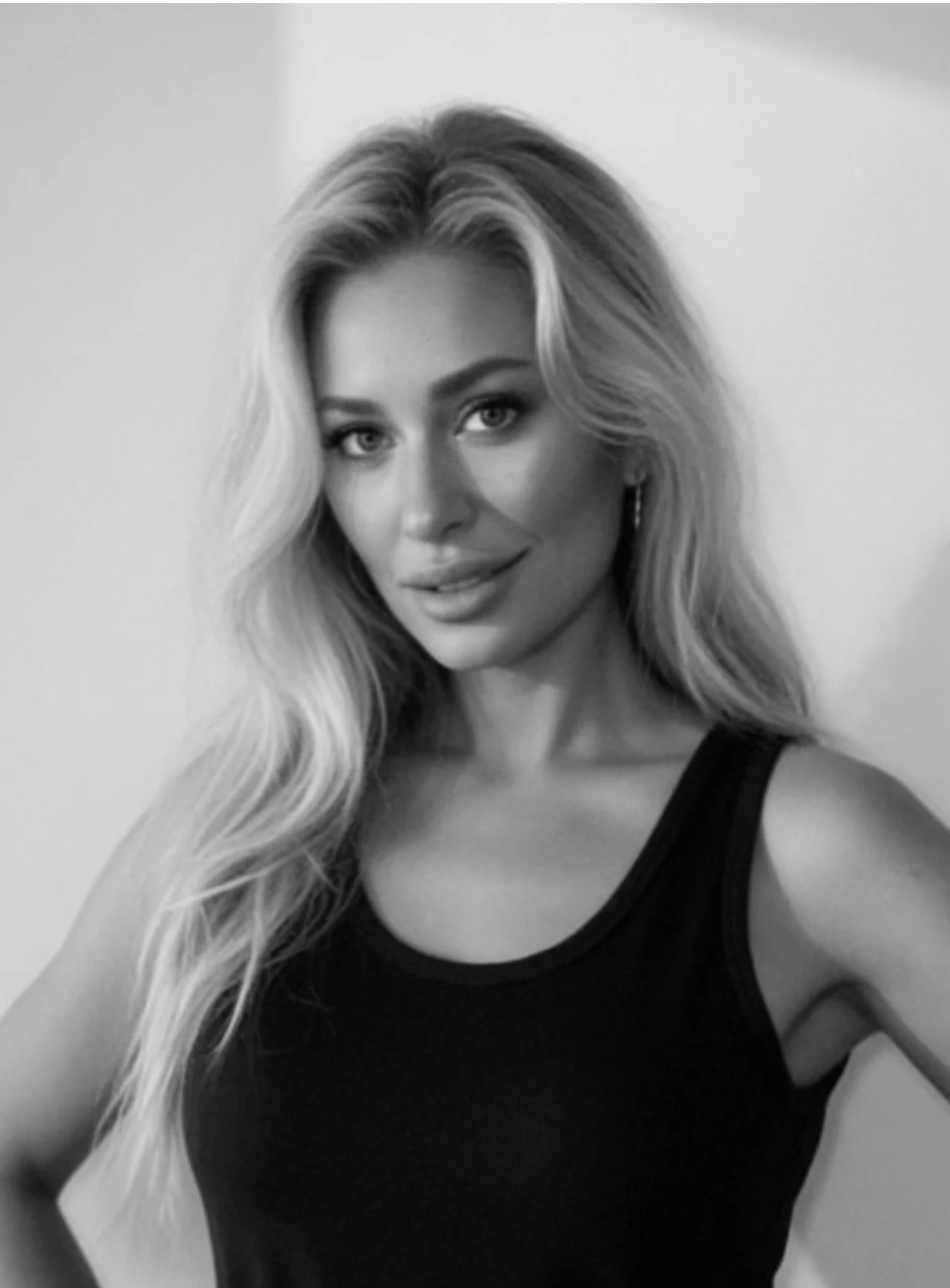 Black and white photo of a smiling woman with long, wavy hair wearing a black tank top, looking at the camera.