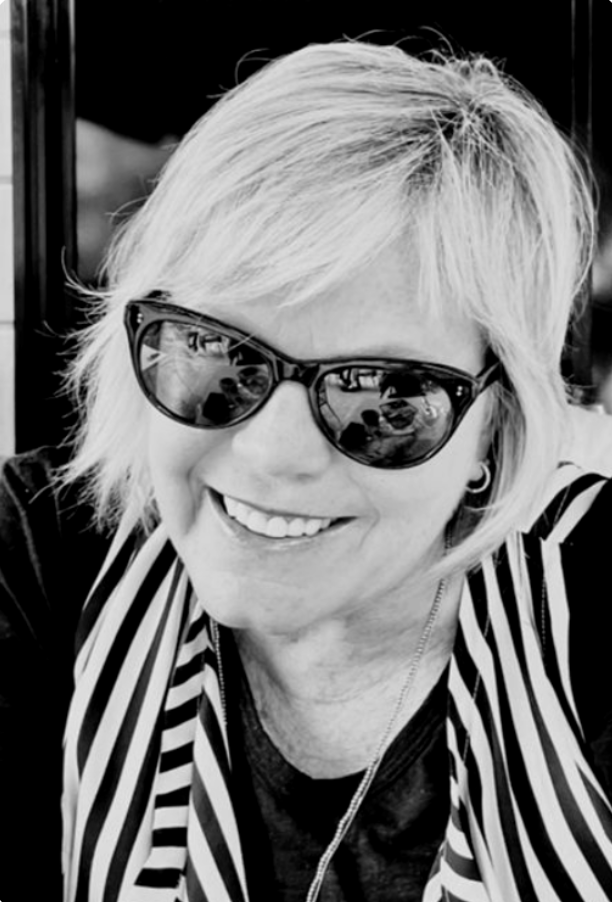 Woman with short blonde hair wearing sunglasses, smiling, dressed in a striped shirt.