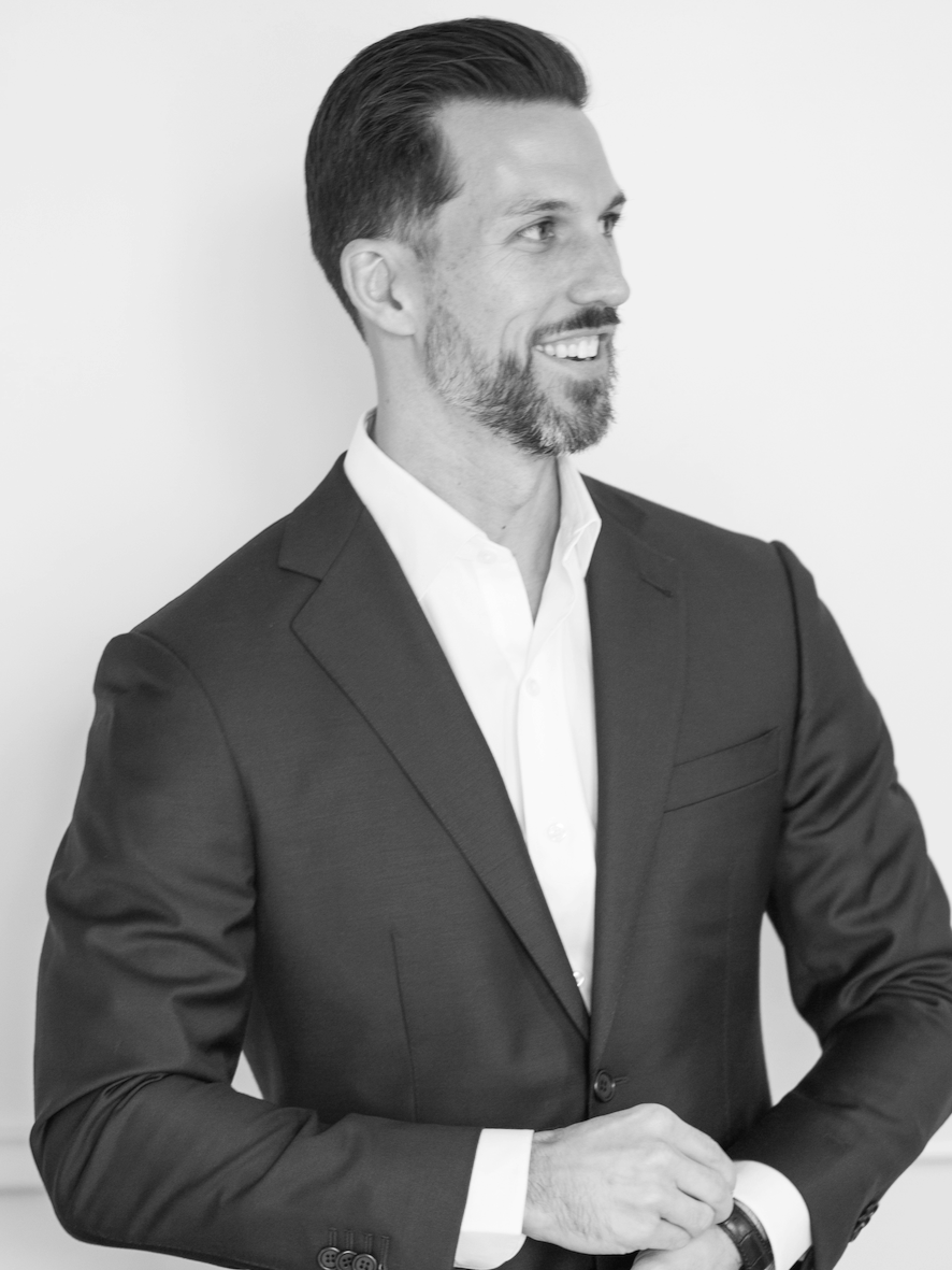 A smiling man with a beard and styled hair wearing a dark suit and white shirt, looking to the right, against a plain light background.