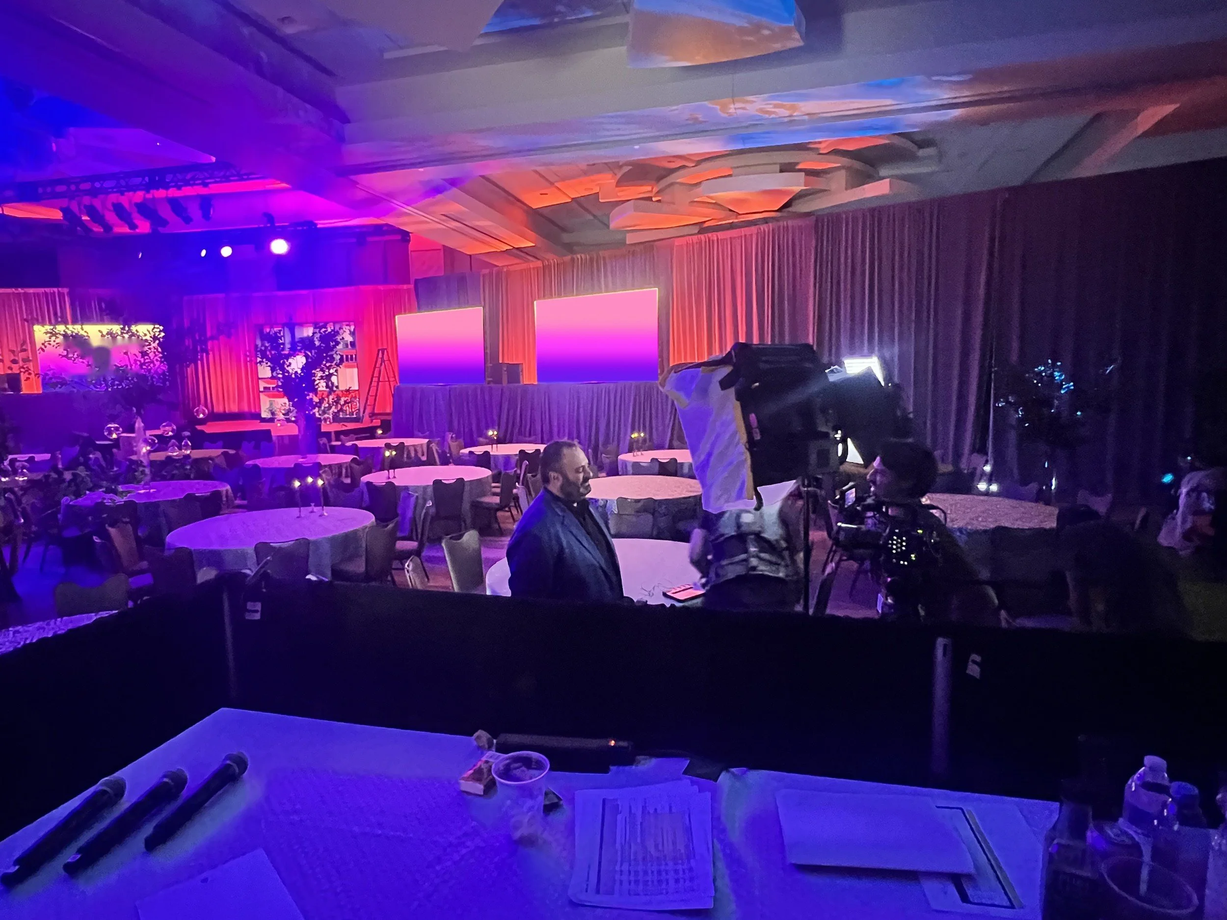 Ballroom dinner: warm red drape, uplit trees, LED screens and a live camera position