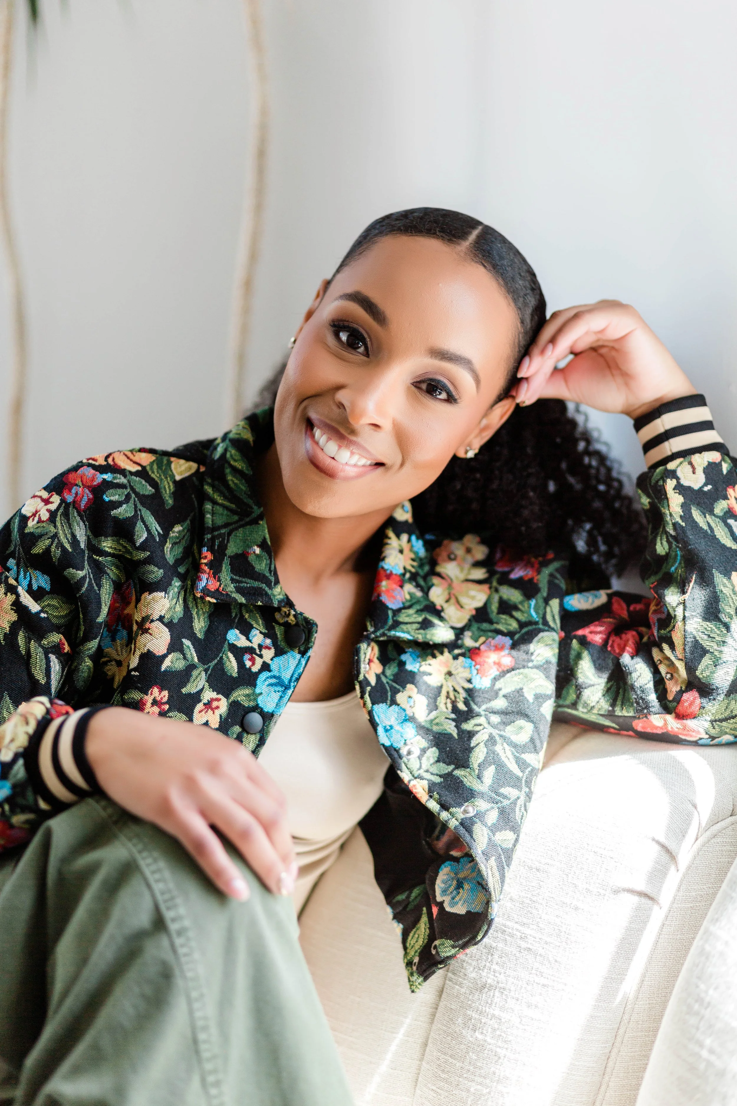 A woman with dark, curly hair, wearing a colorful floral jacket, smiling and resting her head on her hand, sitting on a cream-colored couch with textured fabric.