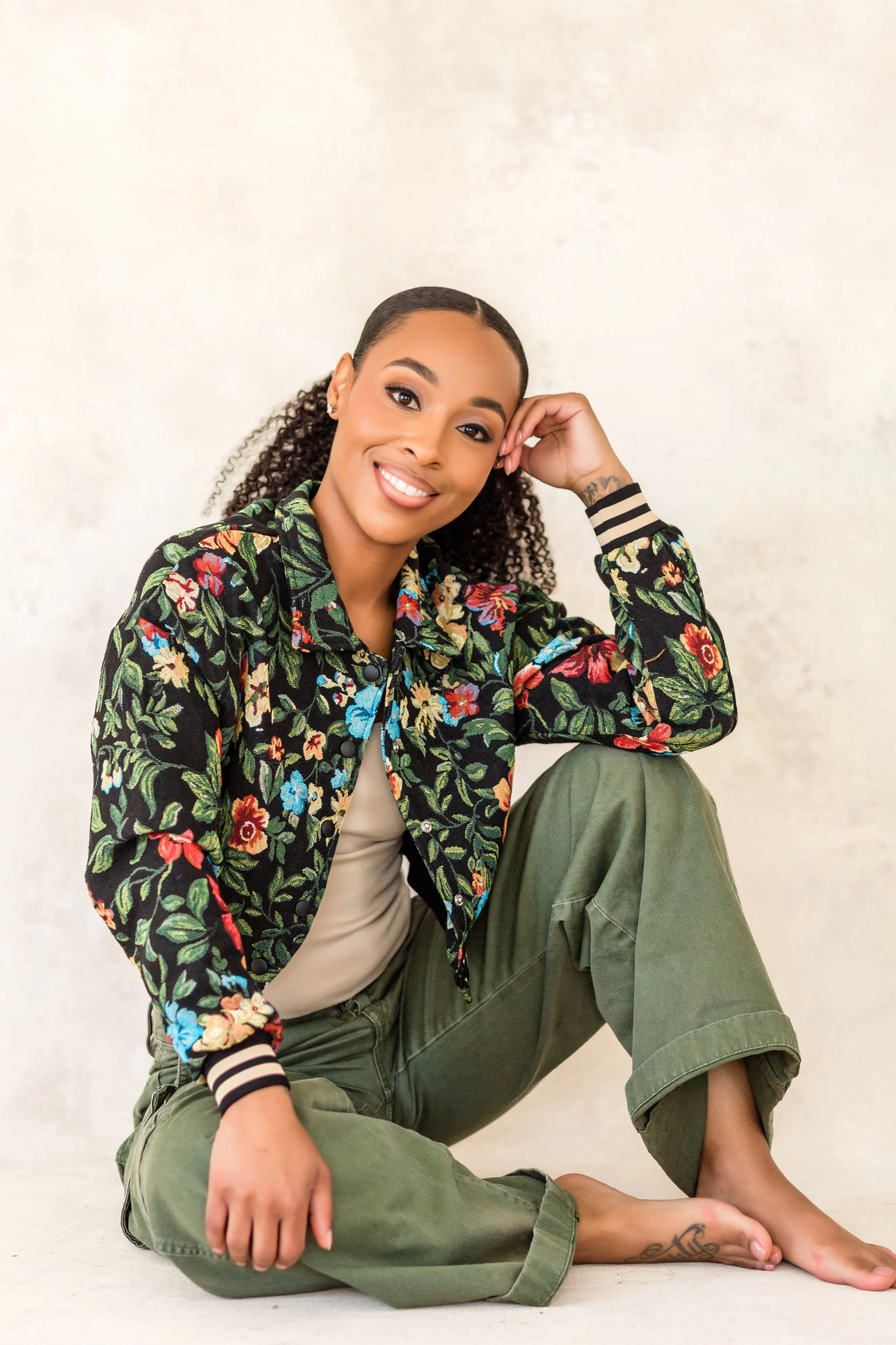A woman with curly hair and tattoos, smiling, sitting on the floor with one knee bent and the other leg extended, wearing a floral jacket, beige shirt, and olive cargo pants.