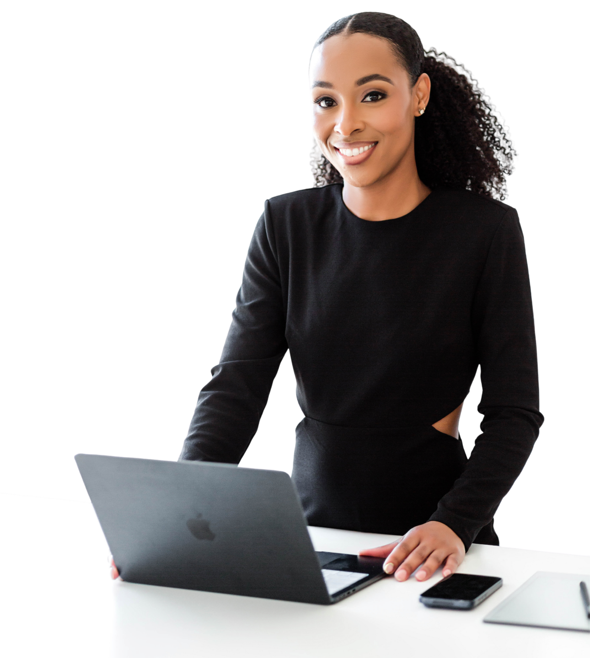 A woman working on a laptop at a white desk with a smartphone nearby, wearing a black top and small earrings, with a black background.