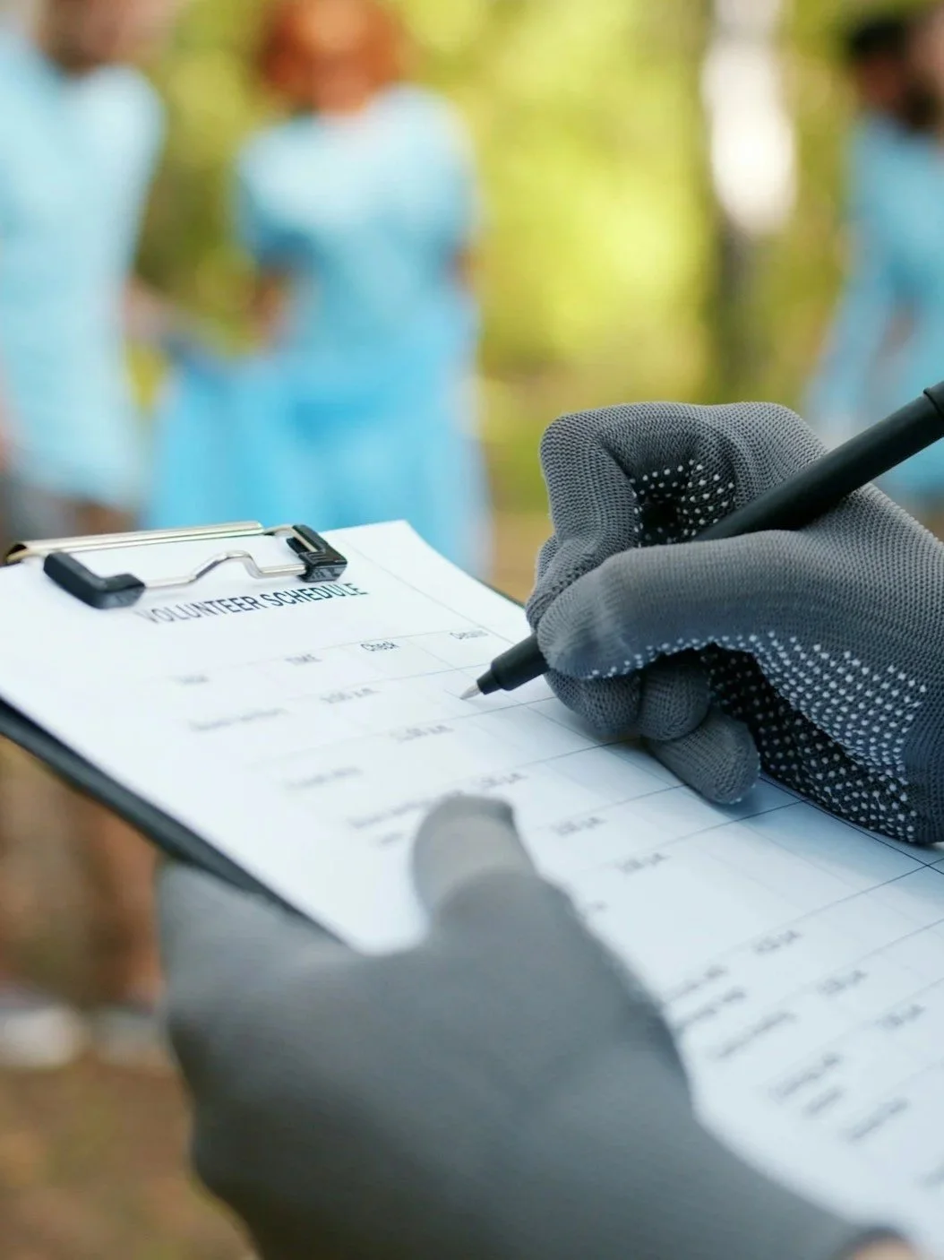 A person wearing gray gloves writes on a volunteer schedule clipboard with a pen, while three people in blue volunteer uniforms stand blurred in the background outdoors.