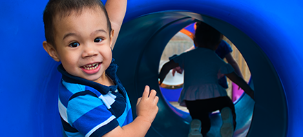 Child smiling and playing inside a blue tunnel with other children in the background.