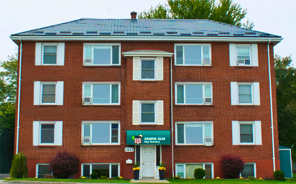 Front view of a red brick three-story building with white window frames, green awning sign for Arbour Glen Day Nursery, and landscaped bushes and flowers in the front yard.