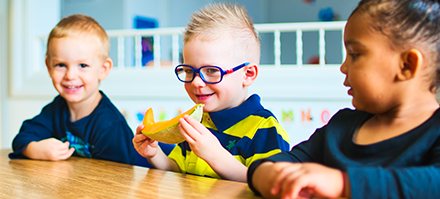 Three children sitting at a table, with one child in the middle eating a banana, smiling, and wearing glasses, in a daycare setting.