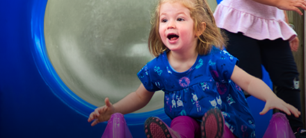 Young girl with blonde hair smiling while sliding down a play tunnel at daycare.