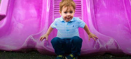 A young boy with light brown hair wearing a light blue shirt and dark pants sitting at the bottom of a pink playground slide.
