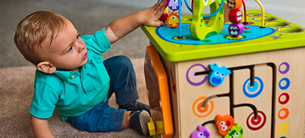 An infant boy in a turquoise shirt and jeans playing with a colorful activity cube on the floor.