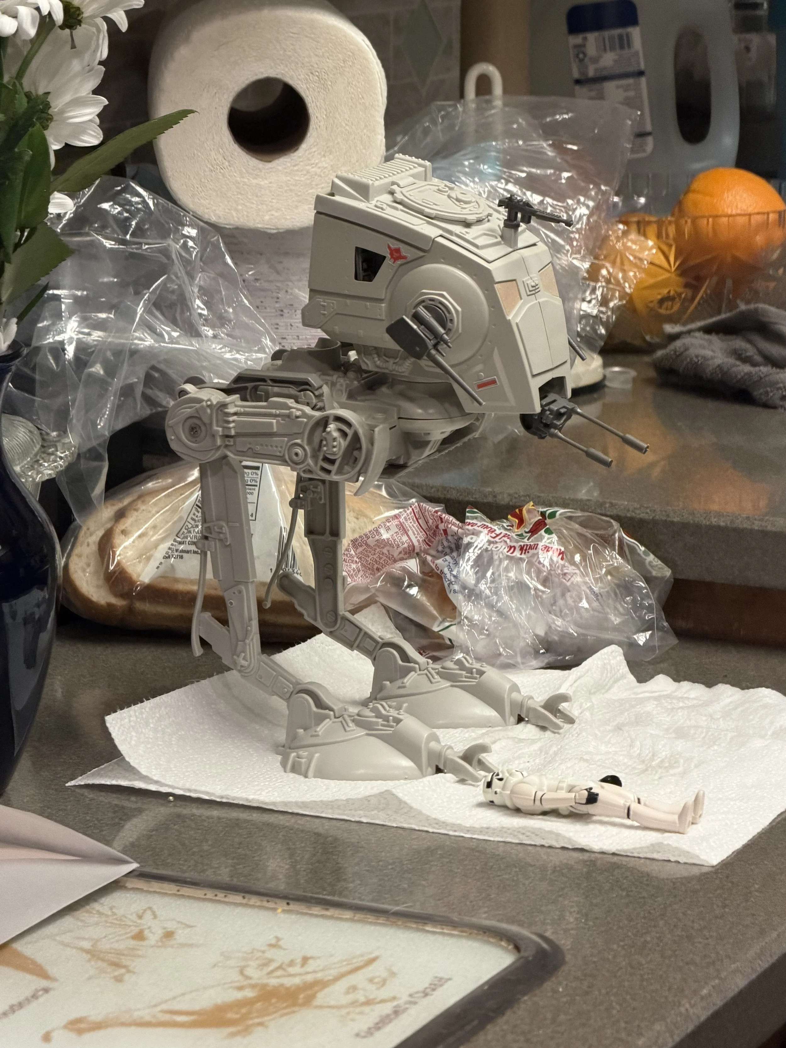 A model of a sci-fi robot or mech, standing on a white paper towel on a kitchen counter. Surrounding objects include a loaf of bread in clear plastic, a roll of paper towels, an orange, and various household items.