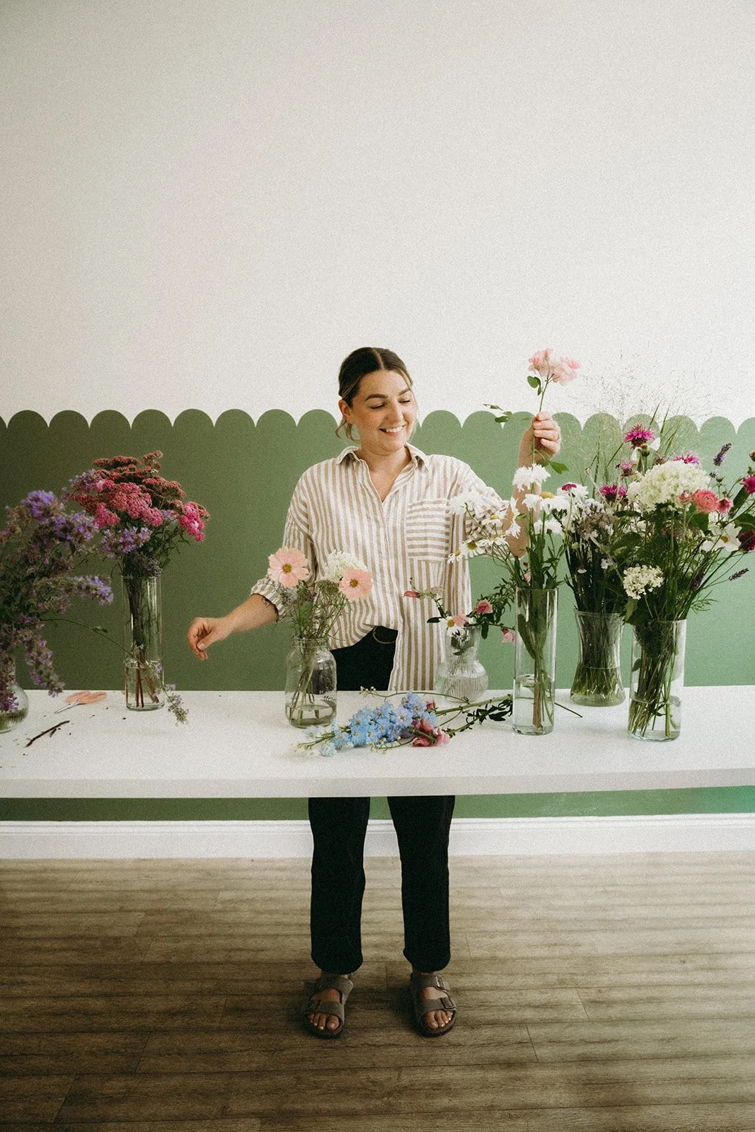 A female florist wearing a striped shirt working with a selection of coloured, garden picked flowers on a workbench in a light studio.