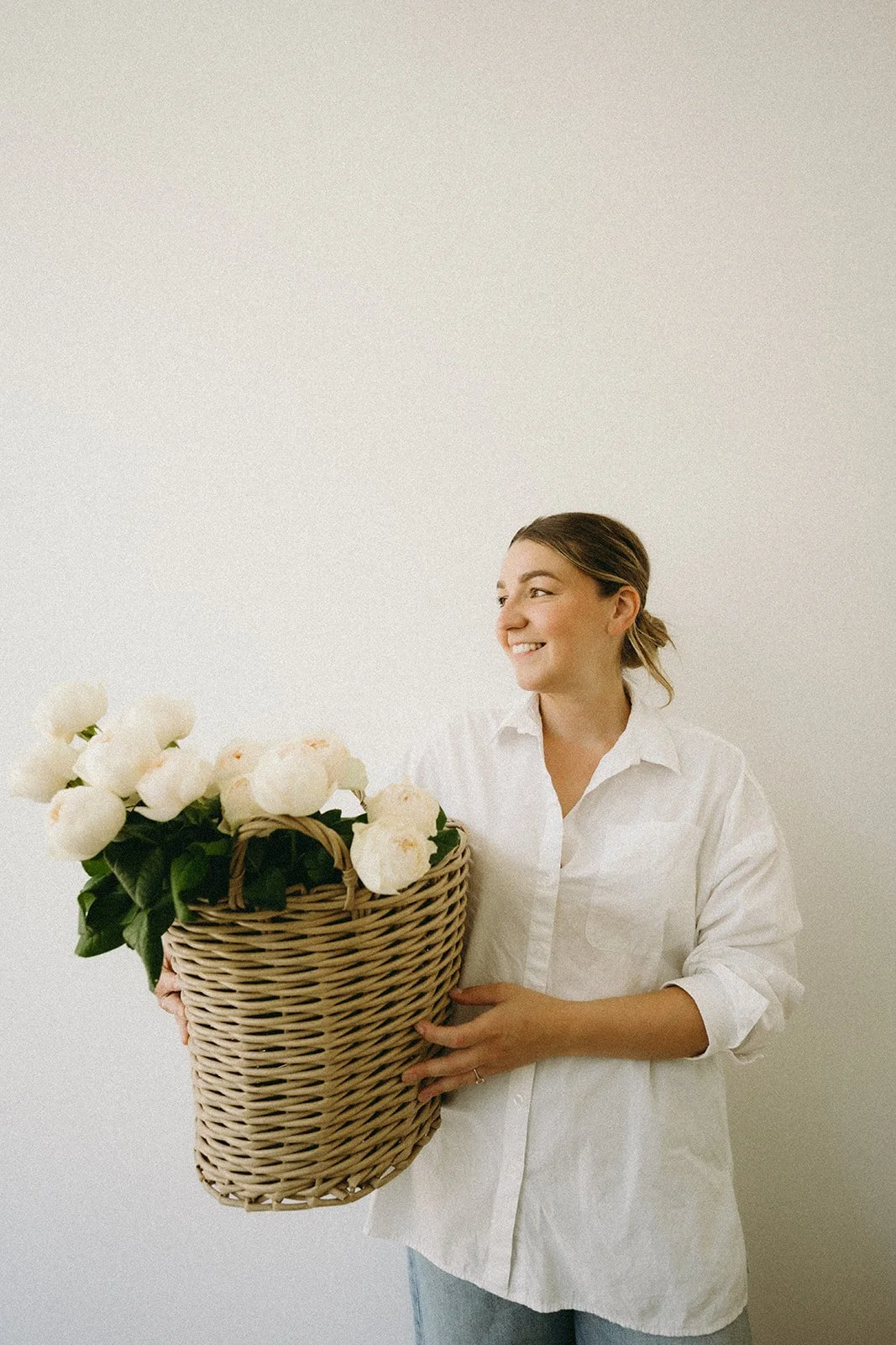 The female owner of Bonnie Blooms holding a rustic basket filled with big, ruffled Roses. Wearing a white shirt and looking into the distance to show a side face profile.