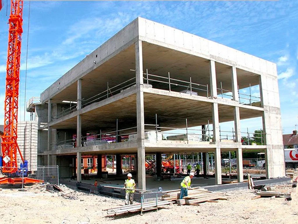 Construction site with a three-story building framework made of concrete, construction workers in safety vests and hard hats, and a tall orange crane on the left, under a blue sky.