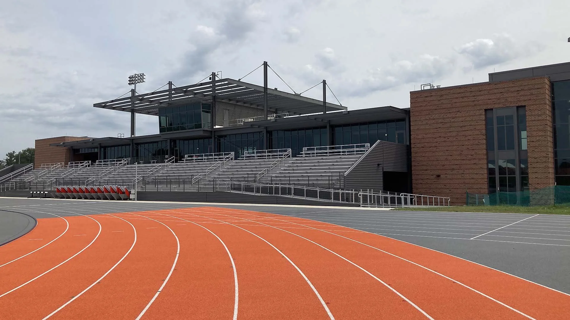 An outdoor track with orange lanes in front of a modern stadium building with glass windows and brick walls. The stadium has tiered seating and a glass control tower.