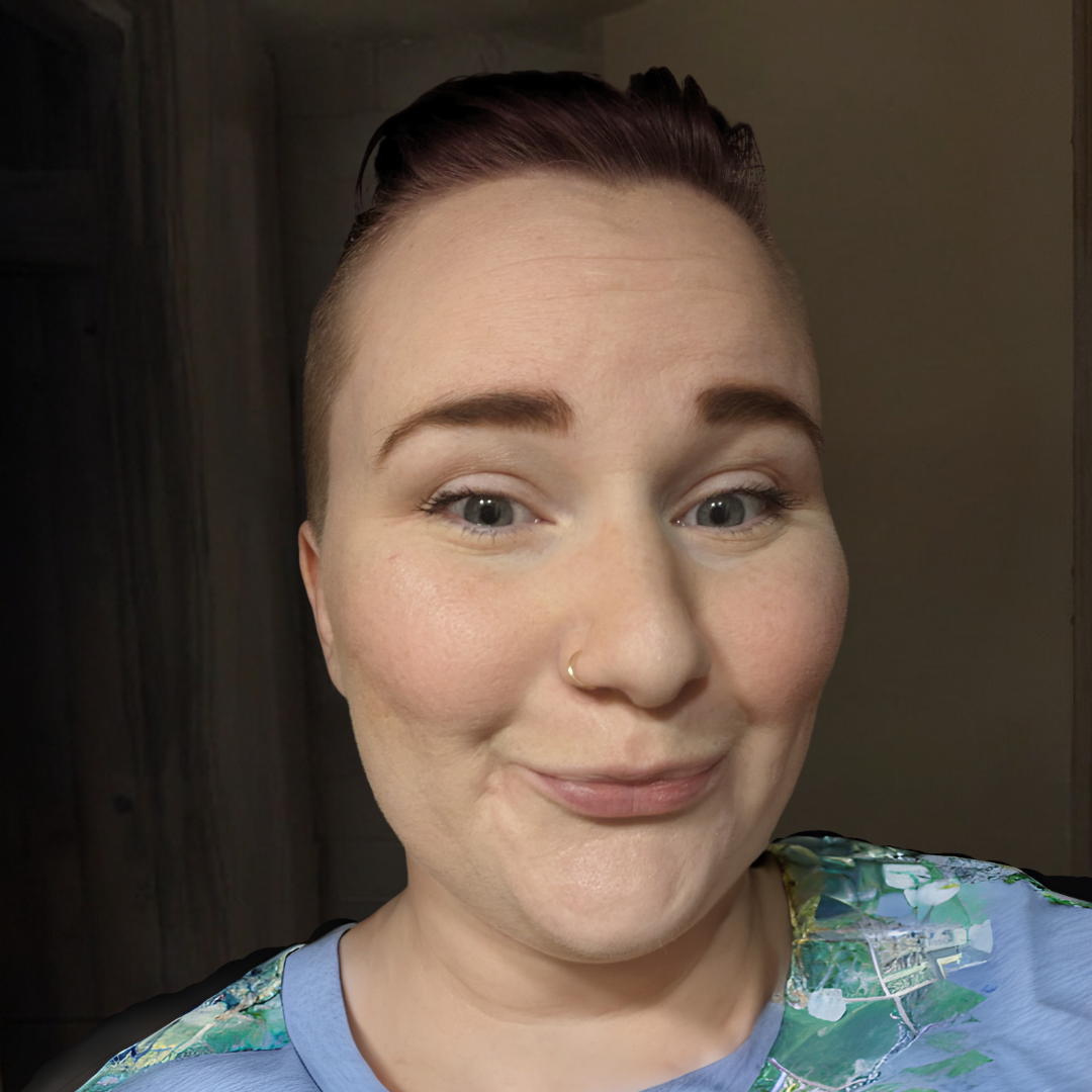 A woman with short brown hair, blue eyes, and a nose ring, smiling and wearing a colorful shirt.