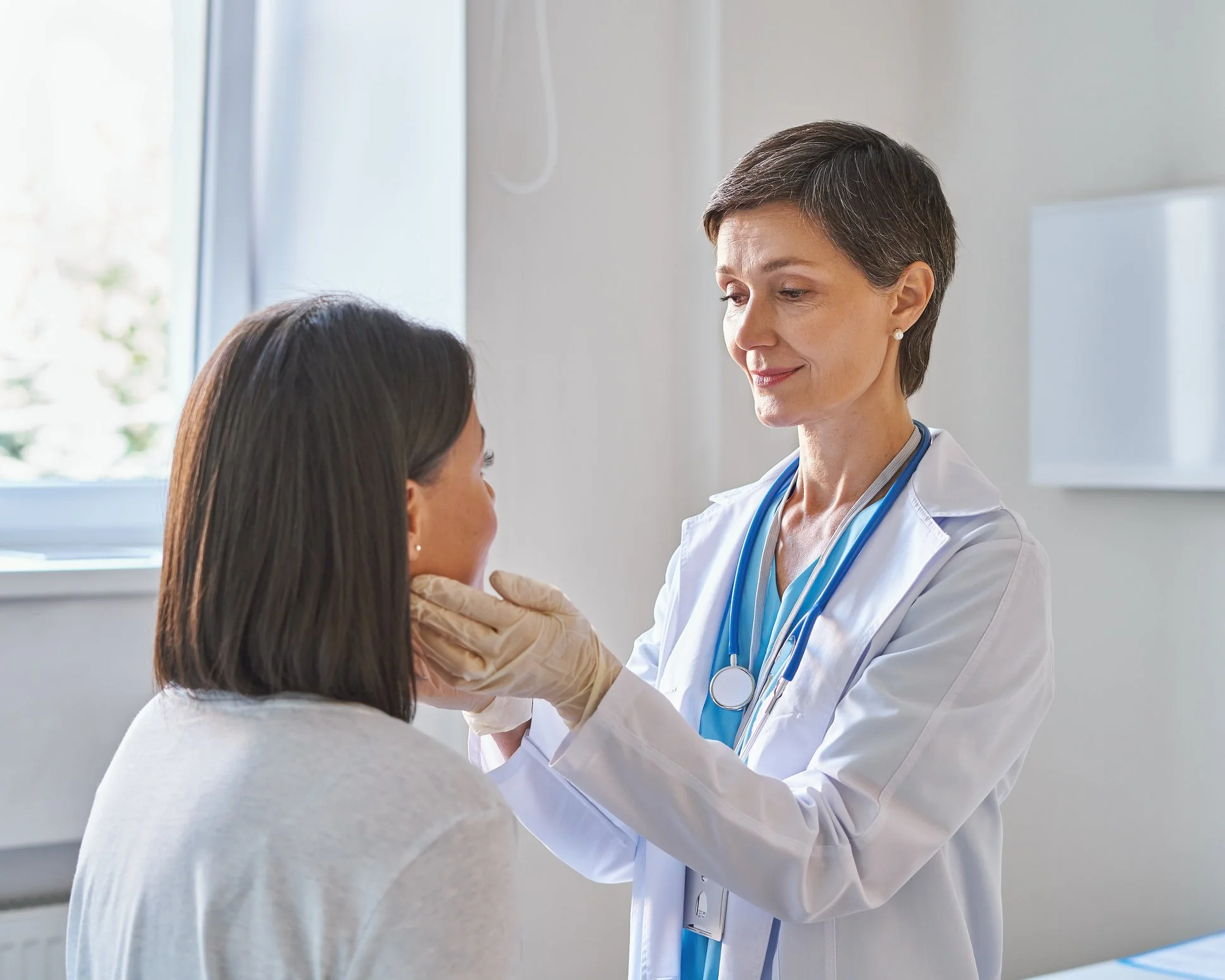 A female doctor examines a patient, a young woman, in a medical office. The doctor has short dark hair, is wearing a white coat and a stethoscope, and smiles gently. The patient has long dark hair and is looking up at the doctor.