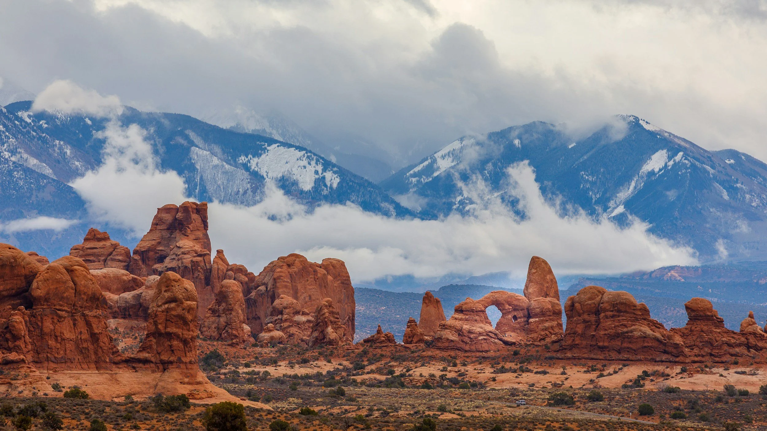 Red rock formations in the foreground with snow-capped mountains and cloudy sky in the background.