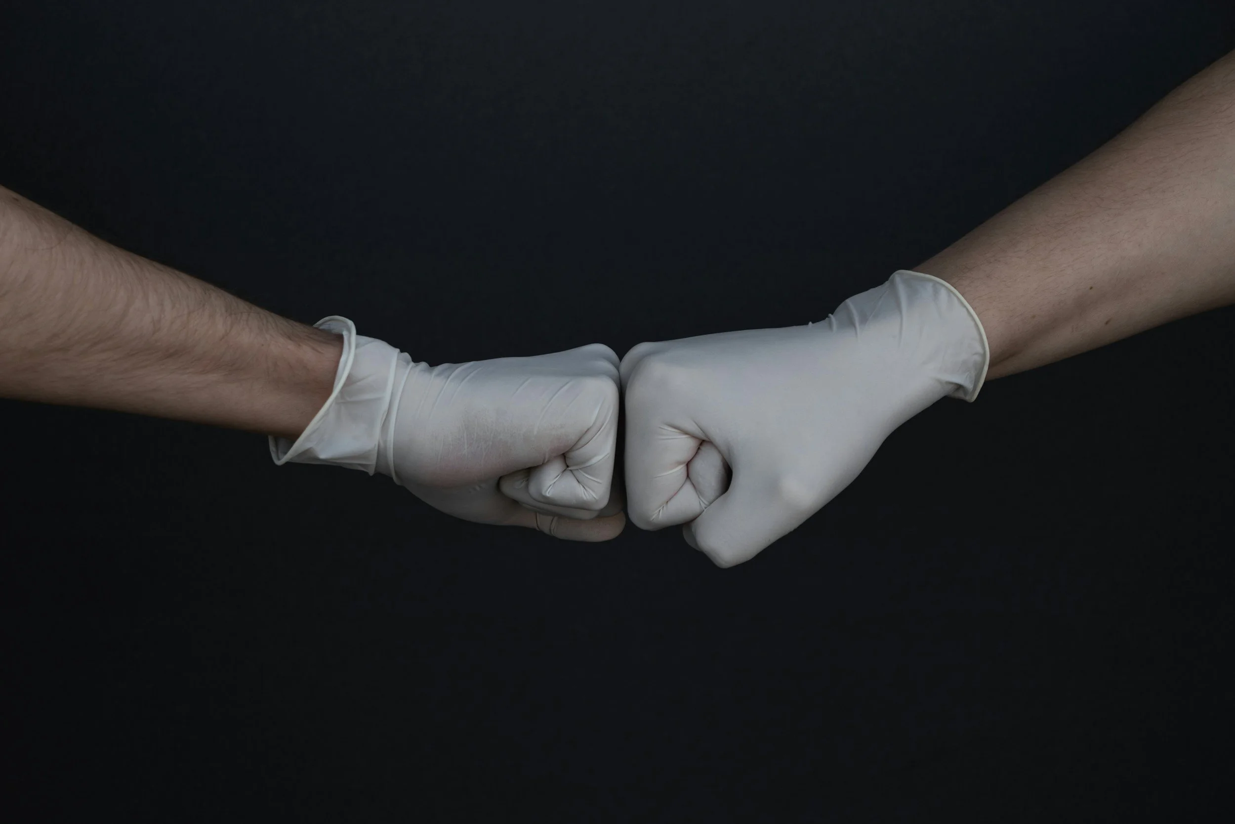 Two arms, one with visible hair, are holding hands against a black background. Both arms are wearing white gloves.