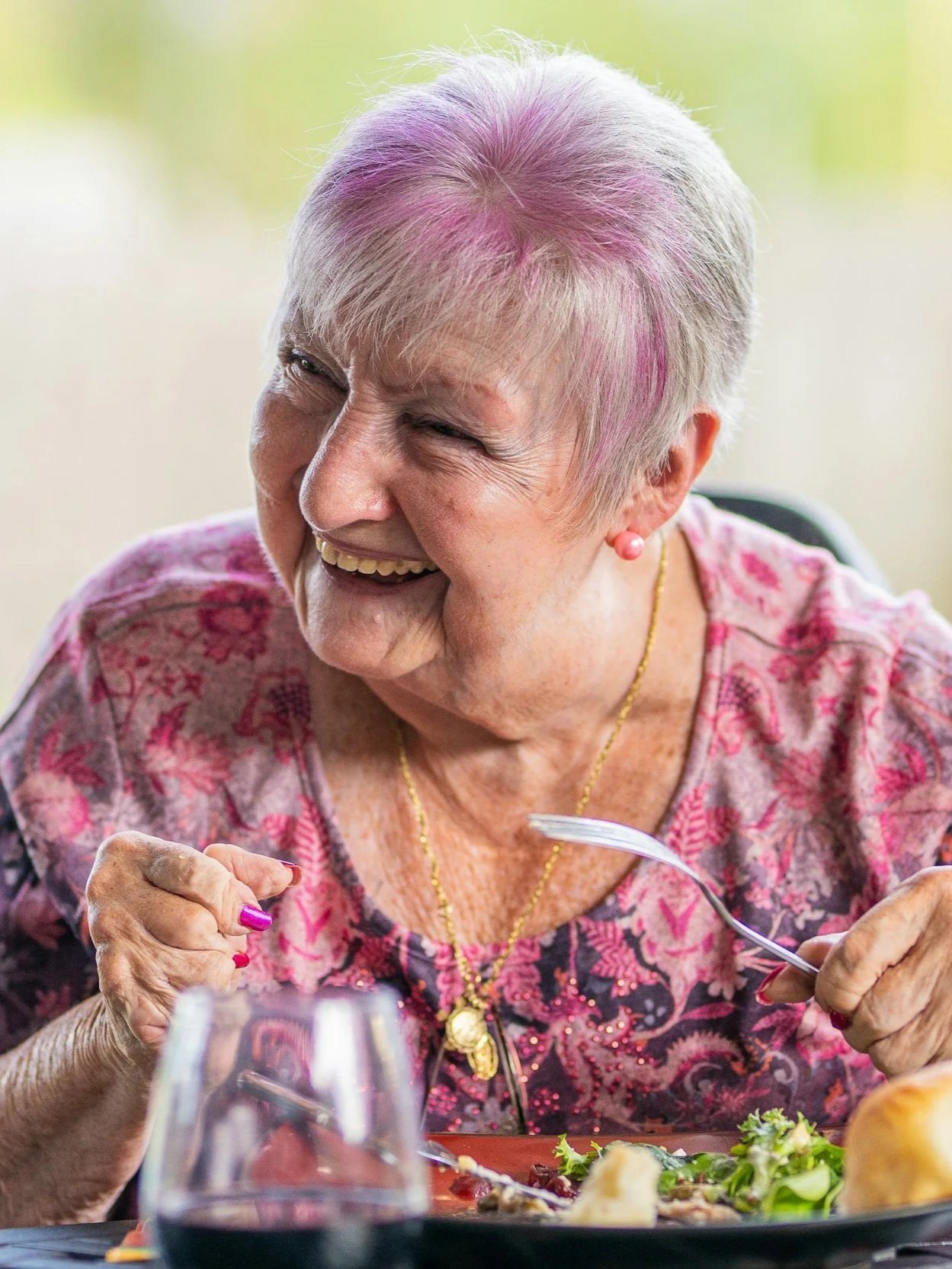 An elderly woman with short gray hair with purple highlights, wearing a floral patterned top, gold jewelry, and pink pearl earrings, is laughing while enjoying a meal at a dining table.
