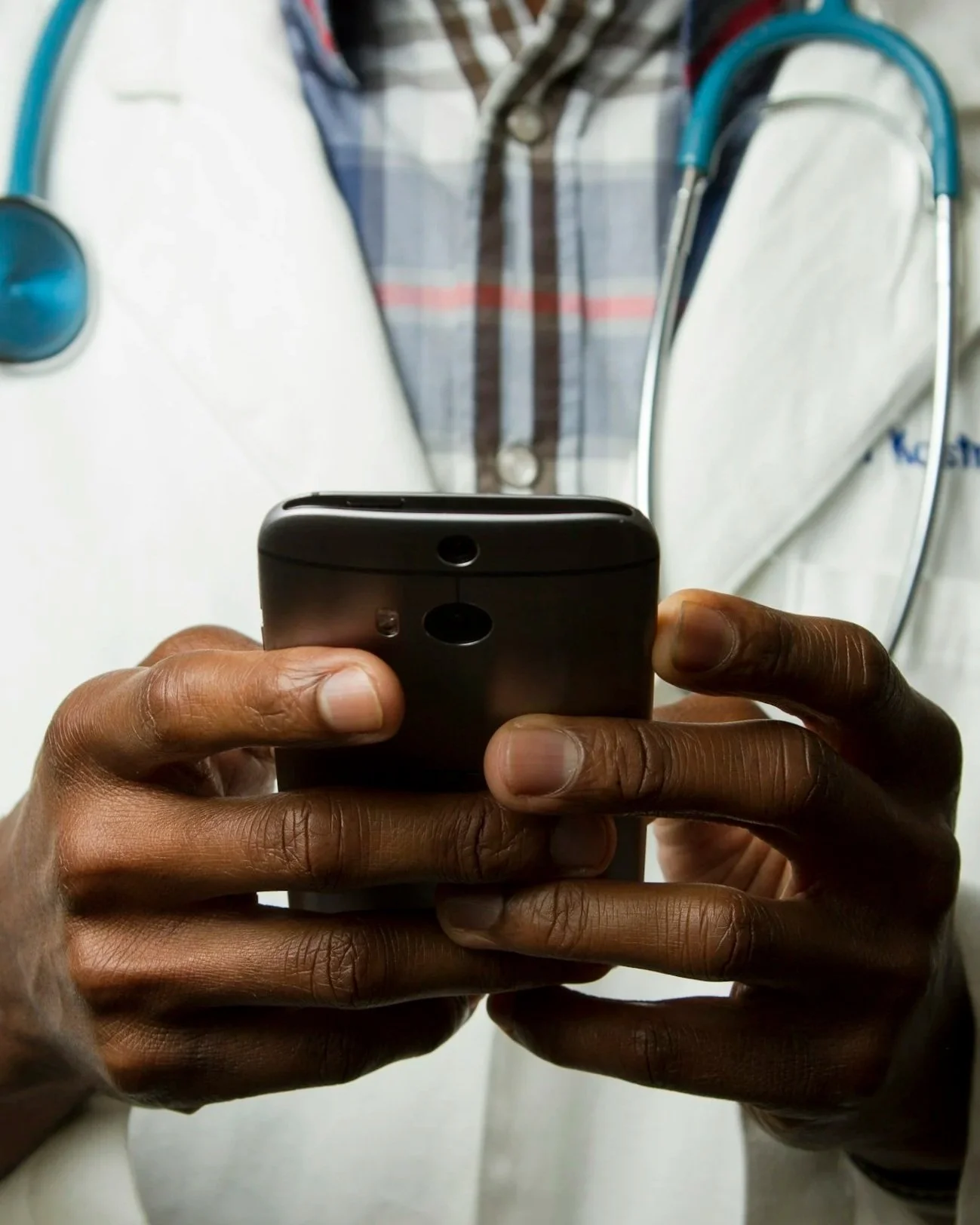 Person using a smartphone with a black case, with another person's hands holding it. A stethoscope and a person wearing a white coat and plaid shirt are visible in the background.