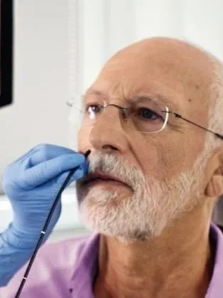 An elderly man wearing glasses and a purple shirt is having a medical nasal swab test administered by a healthcare professional wearing blue gloves.