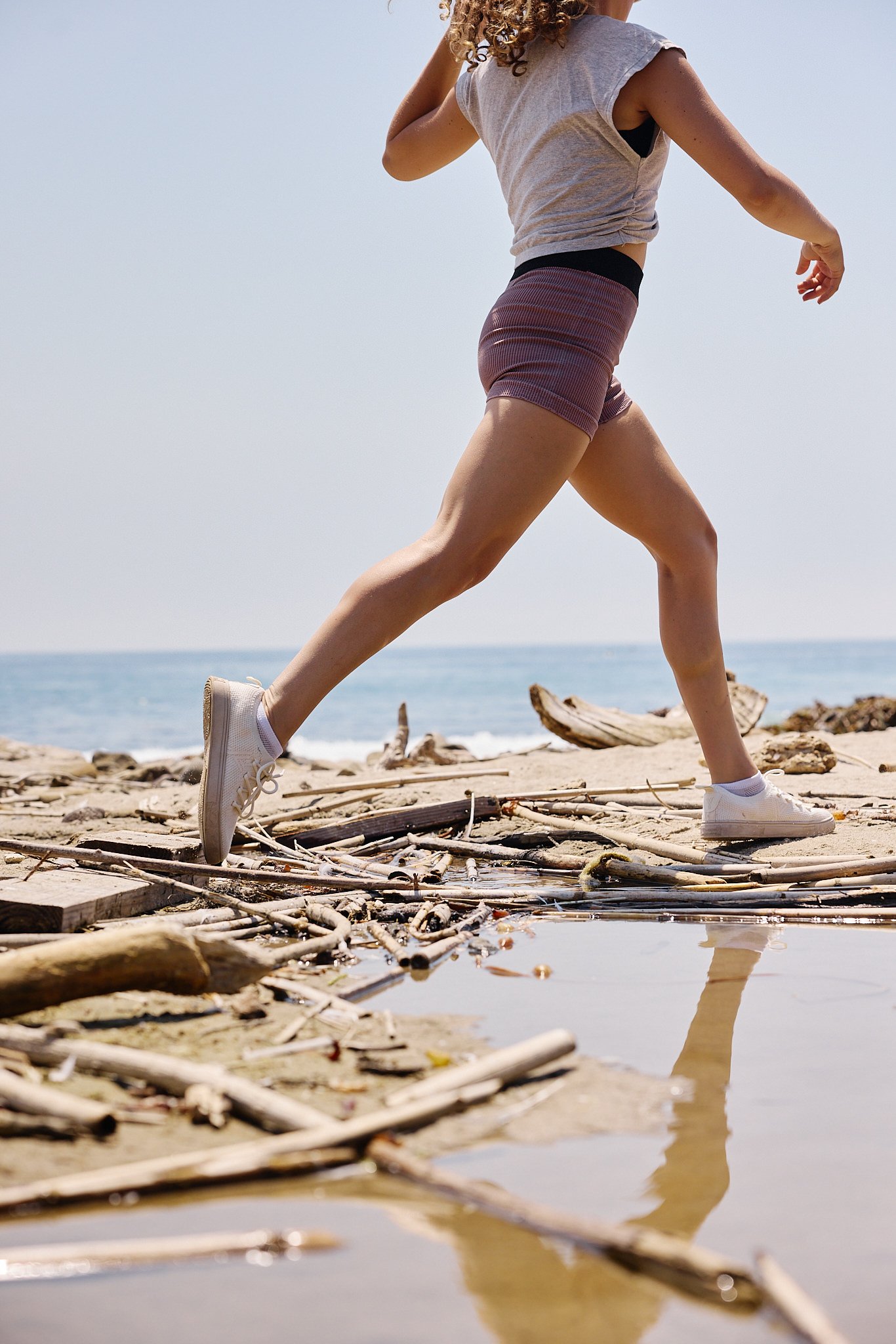 A woman running barefoot on the beach with driftwood and logs scattered on the sand, near shallow water and a cloudy sky.