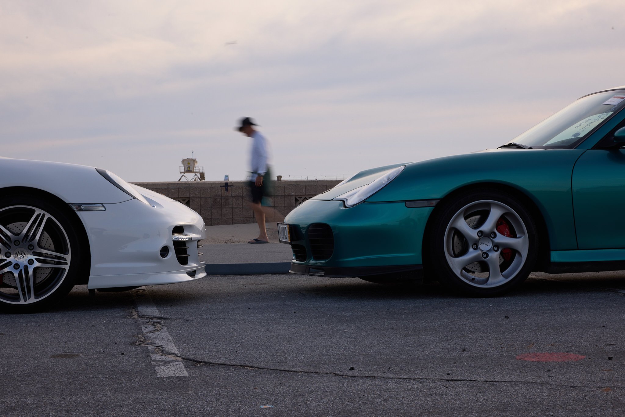 Two sports cars, one white and one teal, parked on a street near a beach promenade with a blurred man walking past and a lifeguard tower in the background under a cloudy sky.