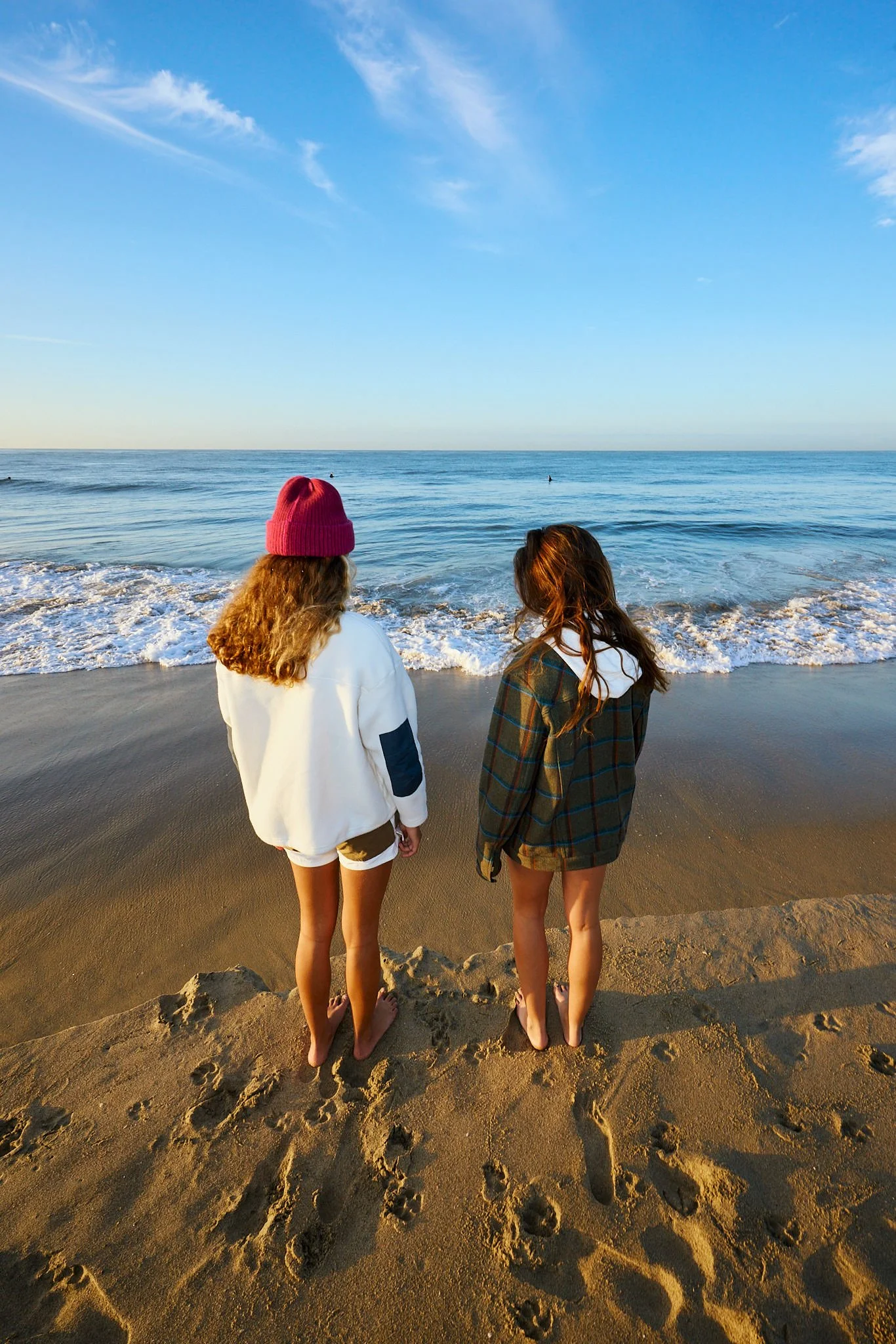 Two women standing on a sandy beach facing the ocean, with clear blue sky and a few clouds above, during sunset or sunrise.